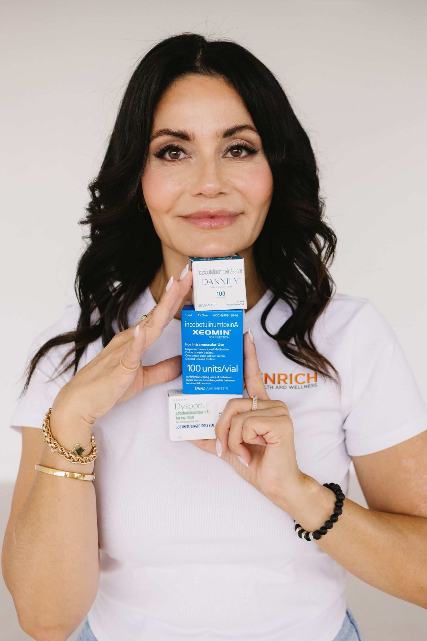 Woman holding Xeomin 100 units botulinum toxin box, wearing Enrich branded white t-shirt and gold jewelry.