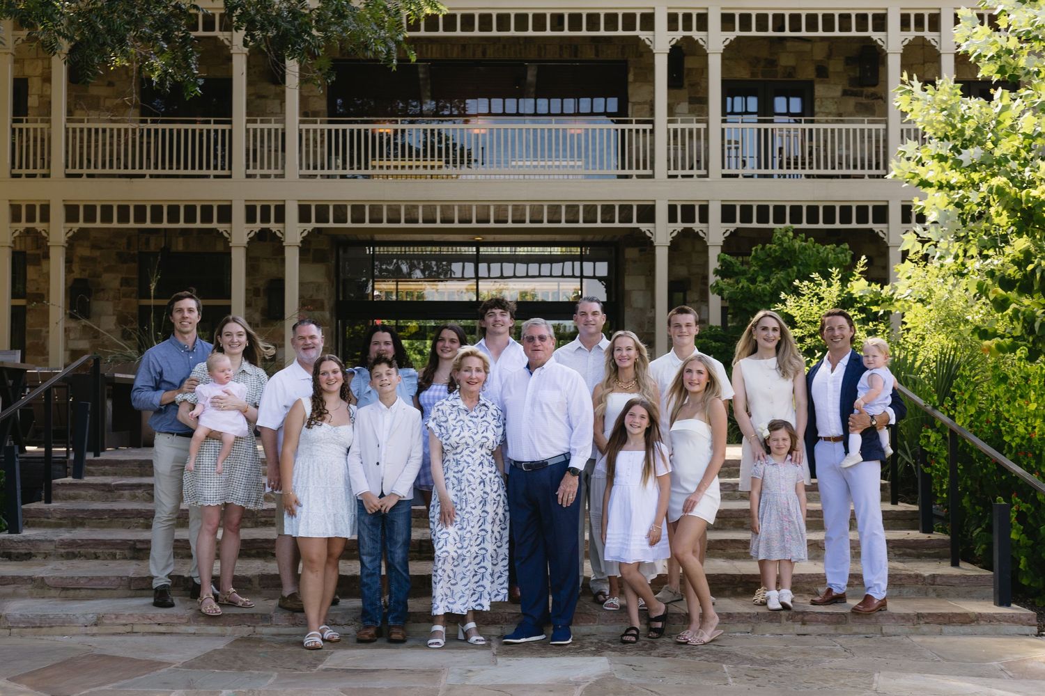 Large family group portrait on stone steps outside elegant two-story building, dressed in coordinating white and neutral outfits.