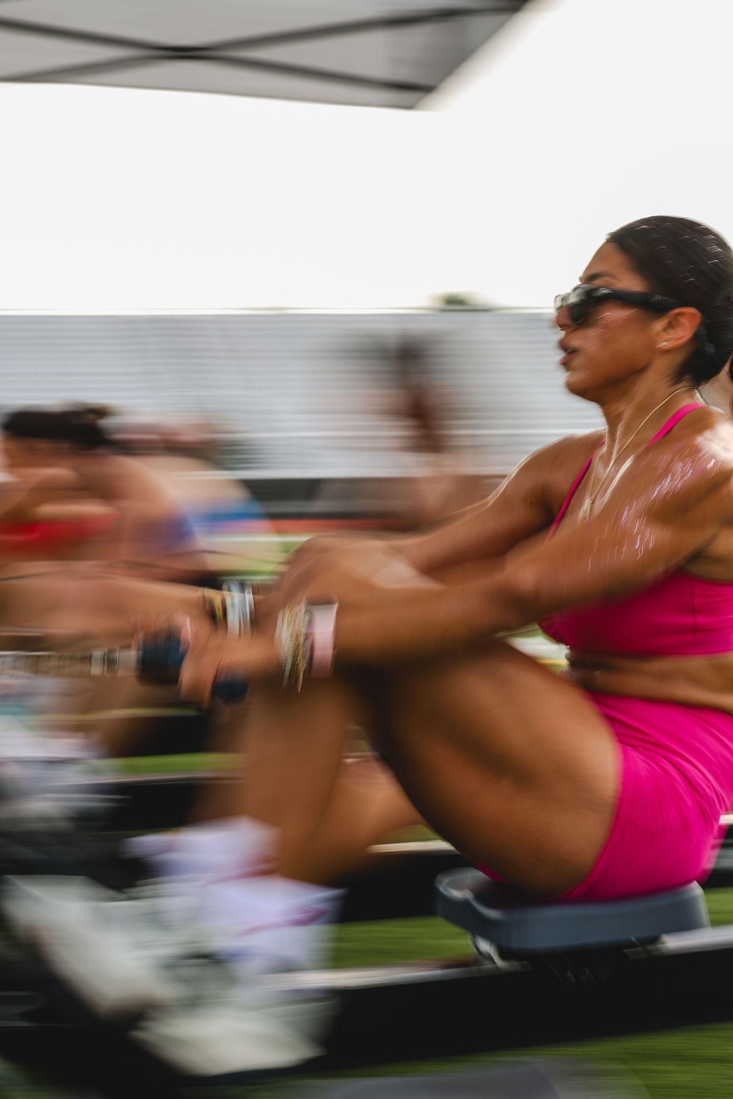 Woman in pink athletic wear rowing on machine outdoors, motion blur capturing intense CrossFit competition effort.