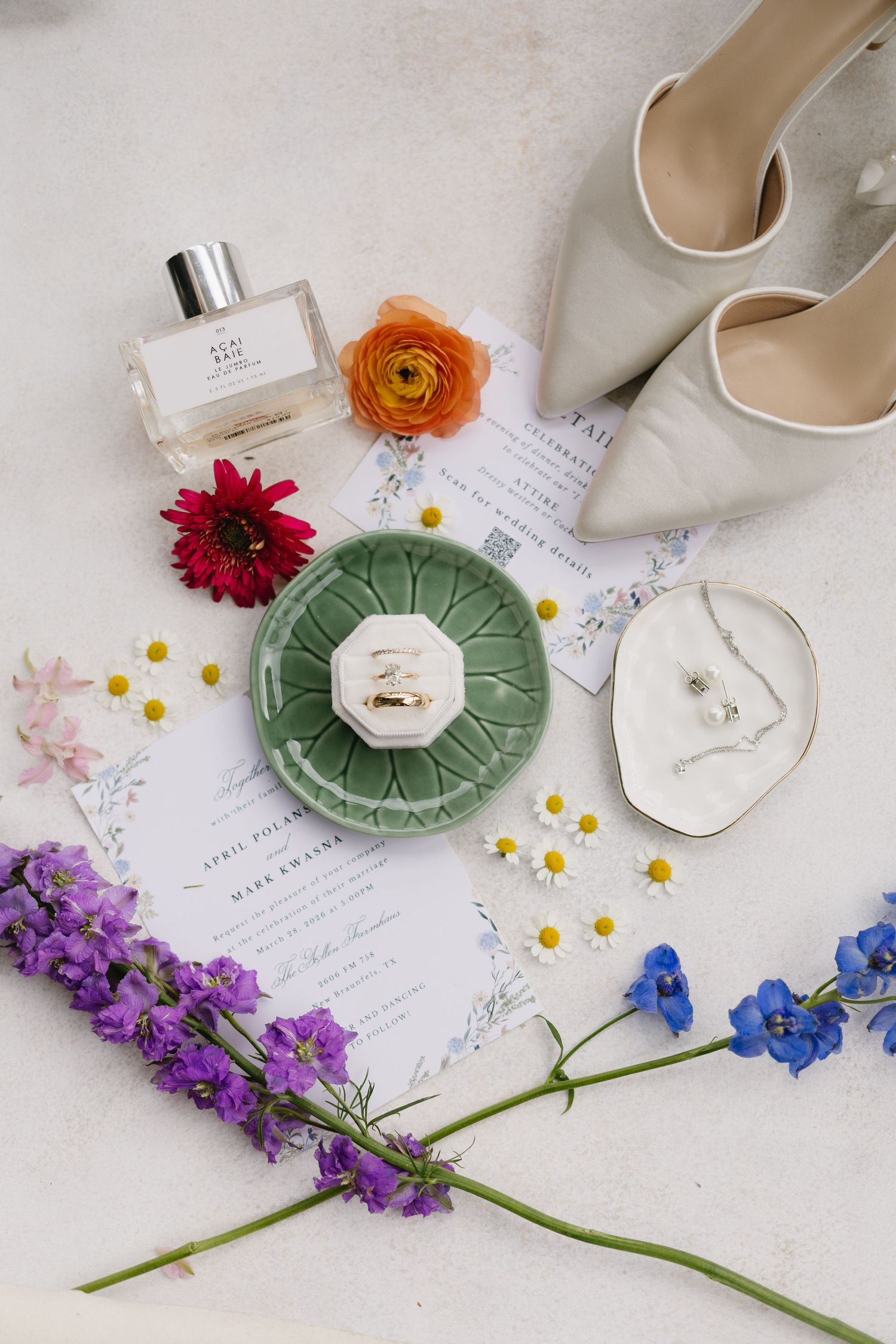 Wedding flat lay with rings on green dish, perfume, heels, florals, and invitation suite on white background.
