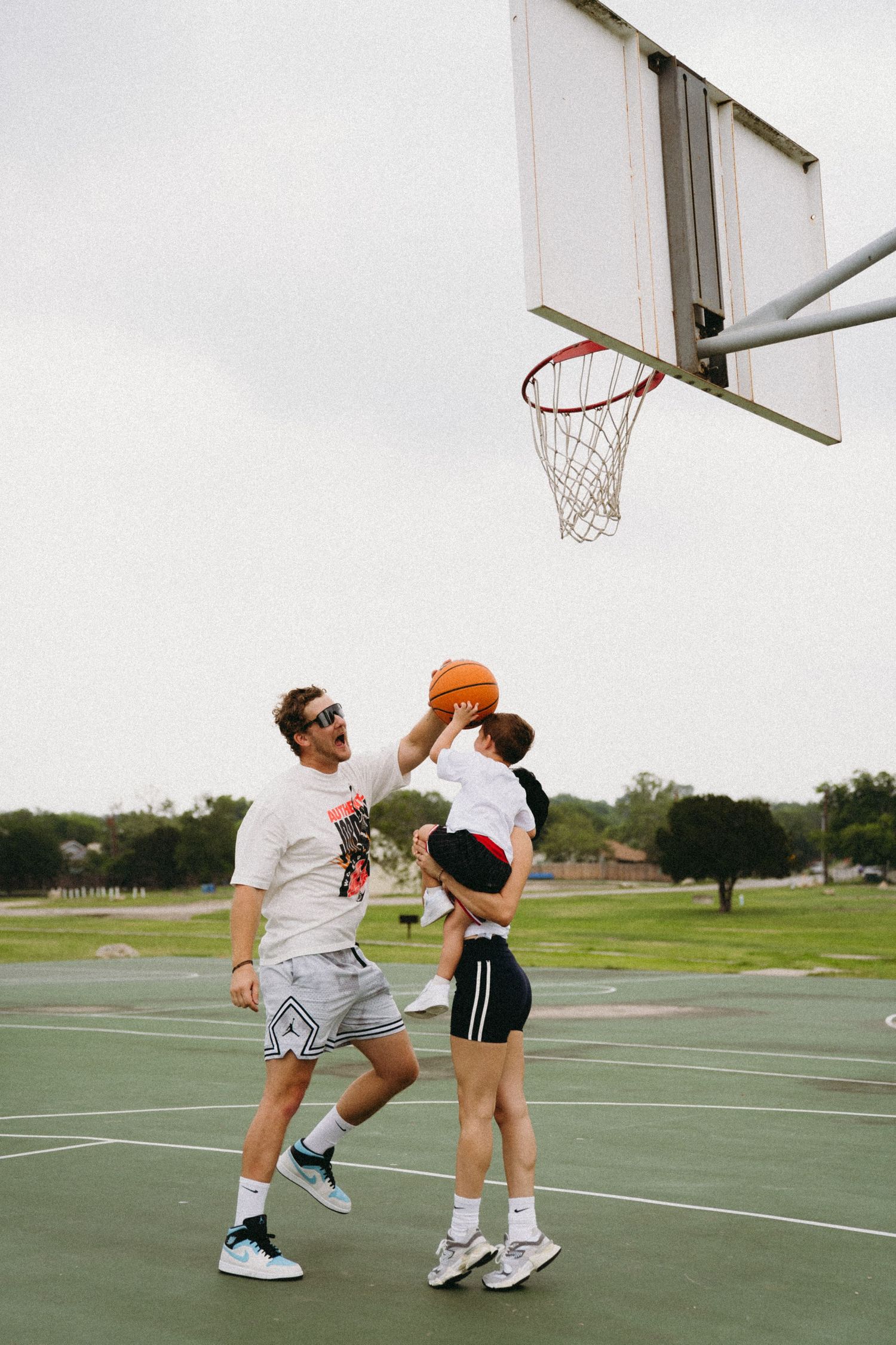 Adult and child playing basketball outdoors, attempting a shot at the hoop on a green court.