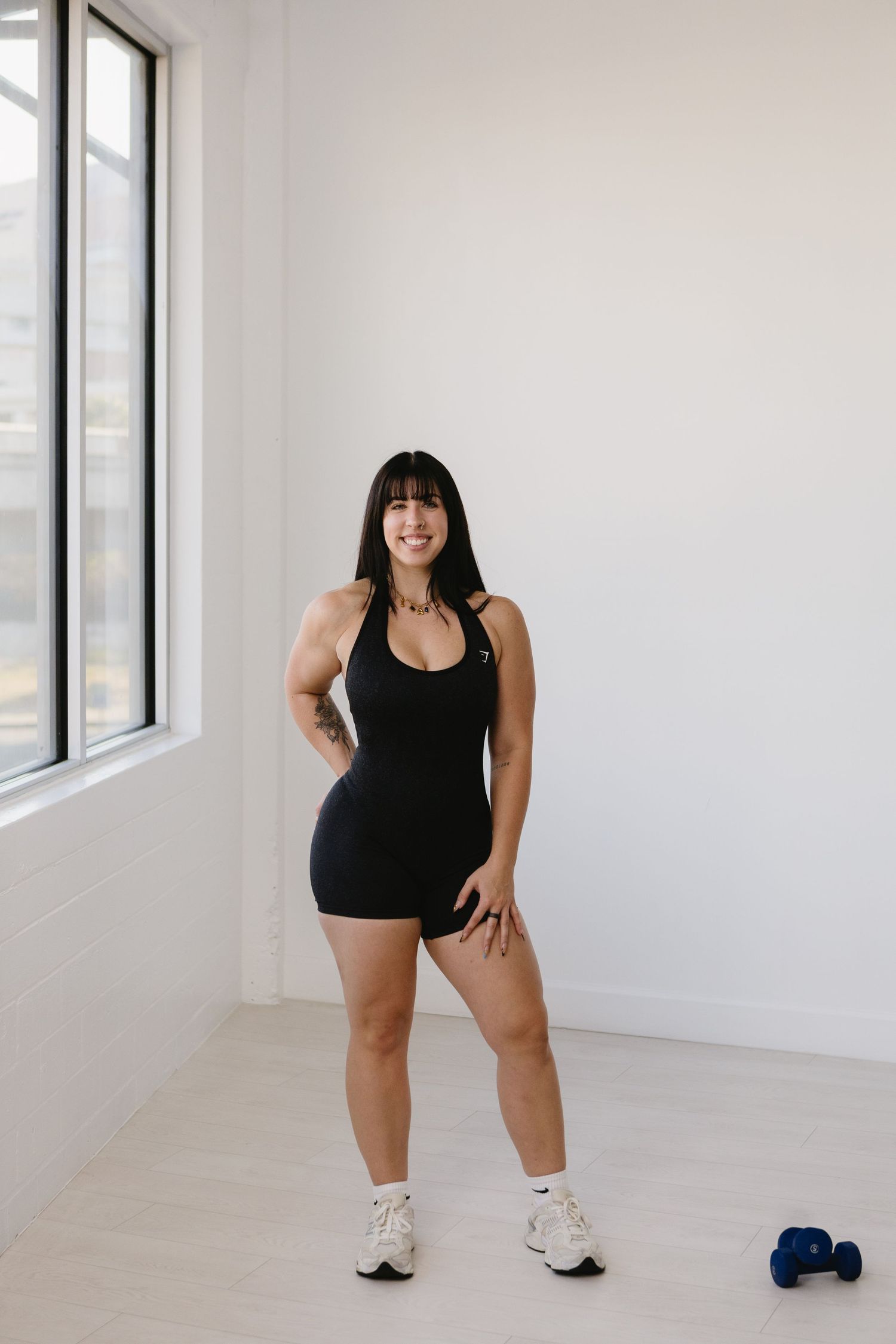 Woman in black athletic romper and white sneakers posing in bright gym with blue dumbbell on floor.
