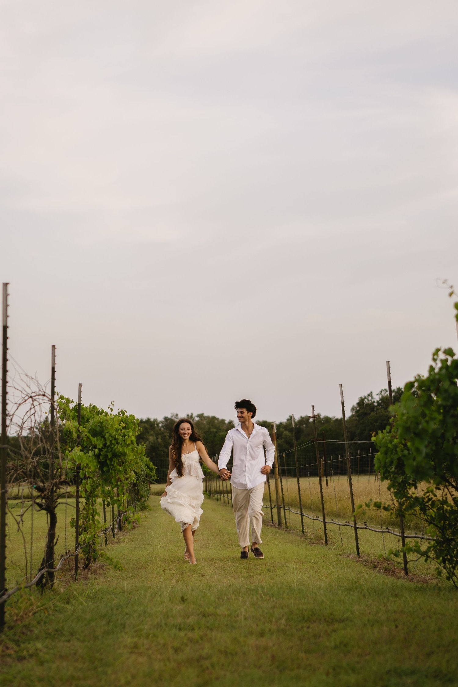 Couple in white attire holding hands and running through lush vineyard rows on their wedding day.