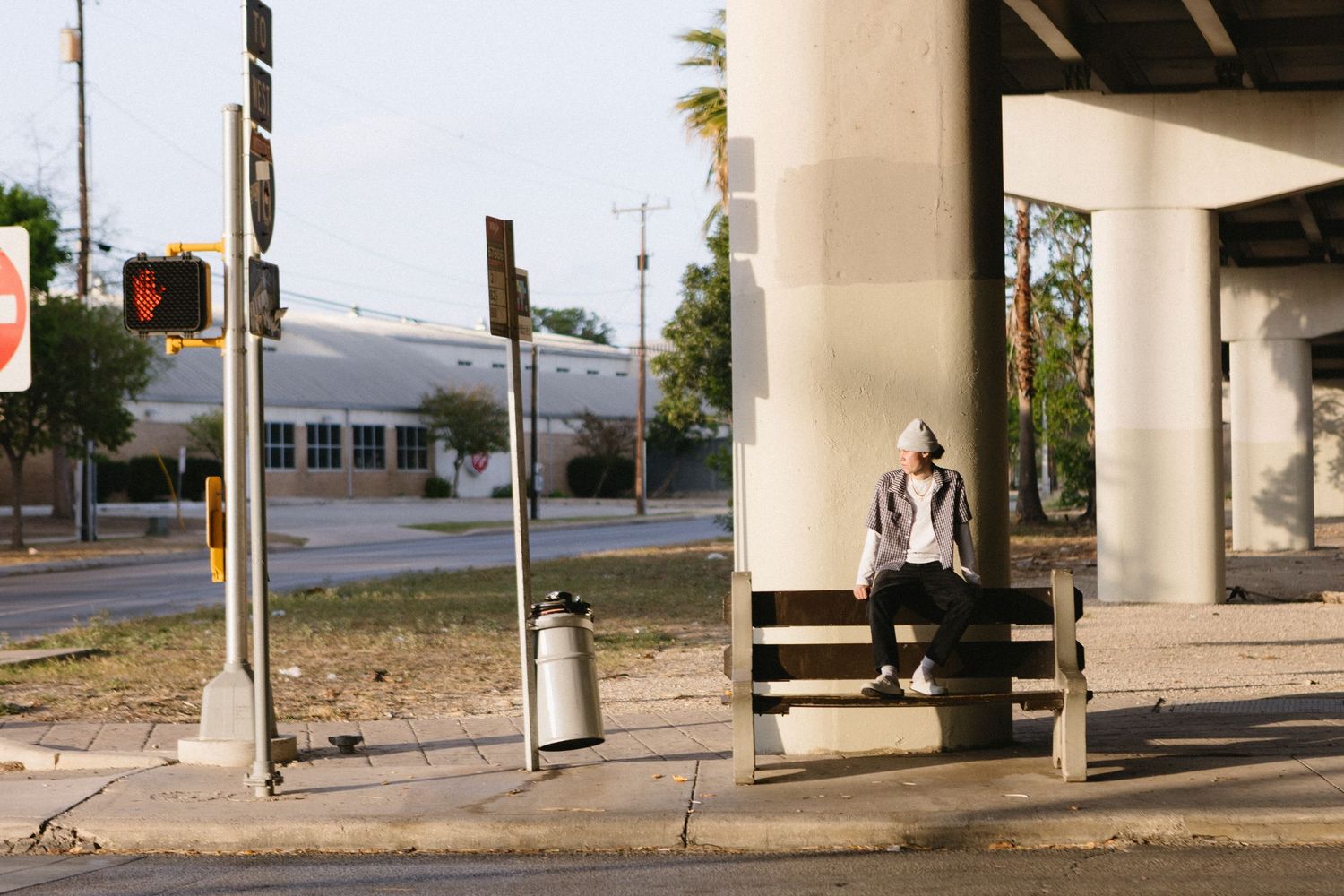 Person sitting alone on a bench under an urban overpass near a city intersection with a trash can nearby.