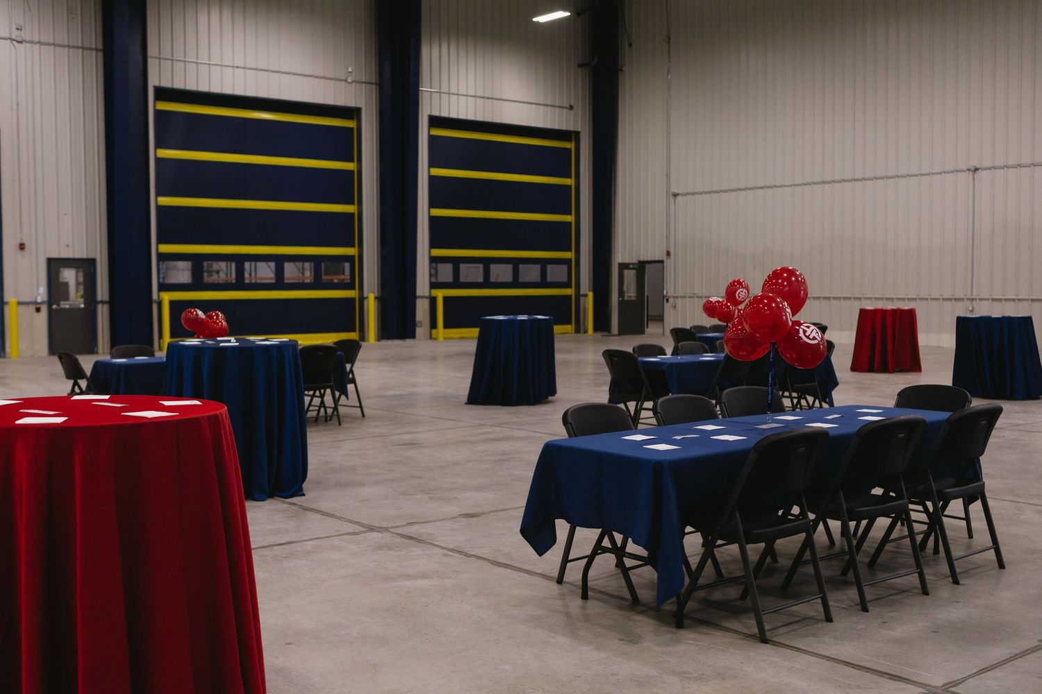 Event space with round tables covered in red and navy blue tablecloths set up in front of industrial loading dock doors.