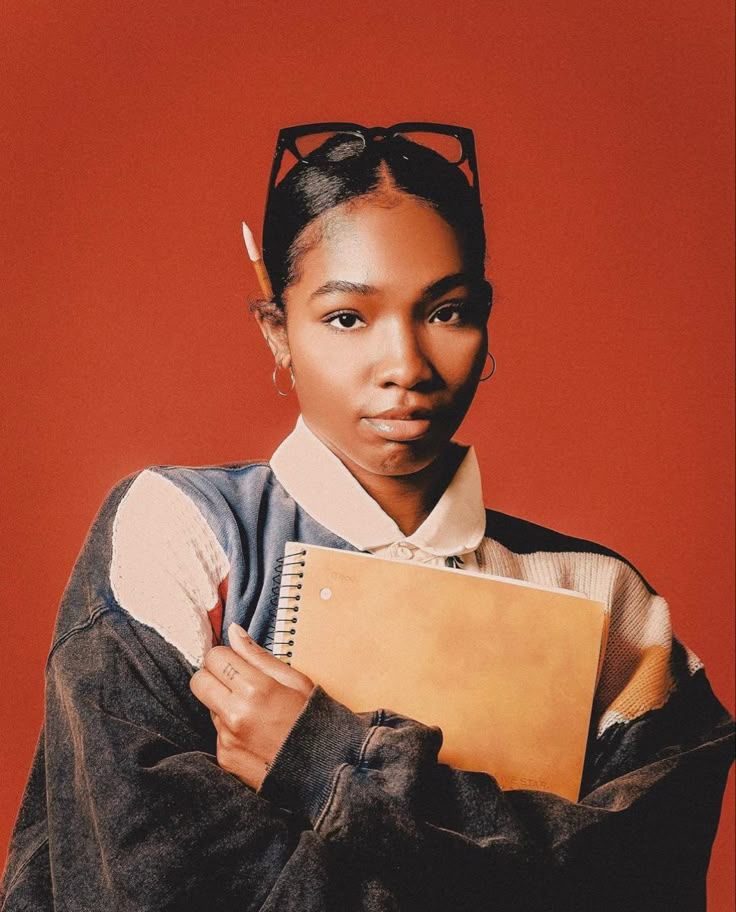 Stylish student wearing sunglasses on head holds a notebook against vibrant orange backdrop.