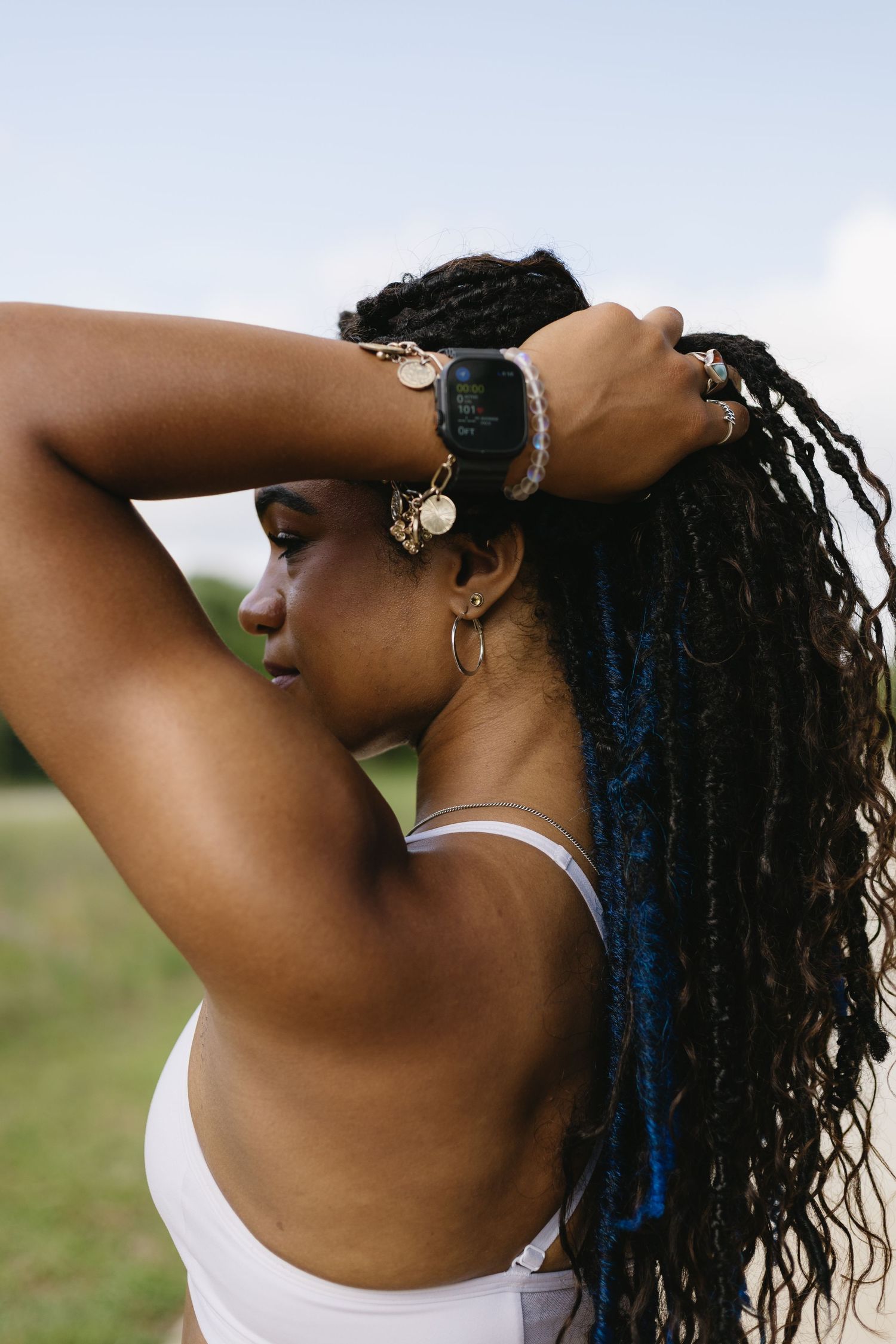 A person with long braided hair and jewelry tilts their head back outdoors against a green background in the sunlight.