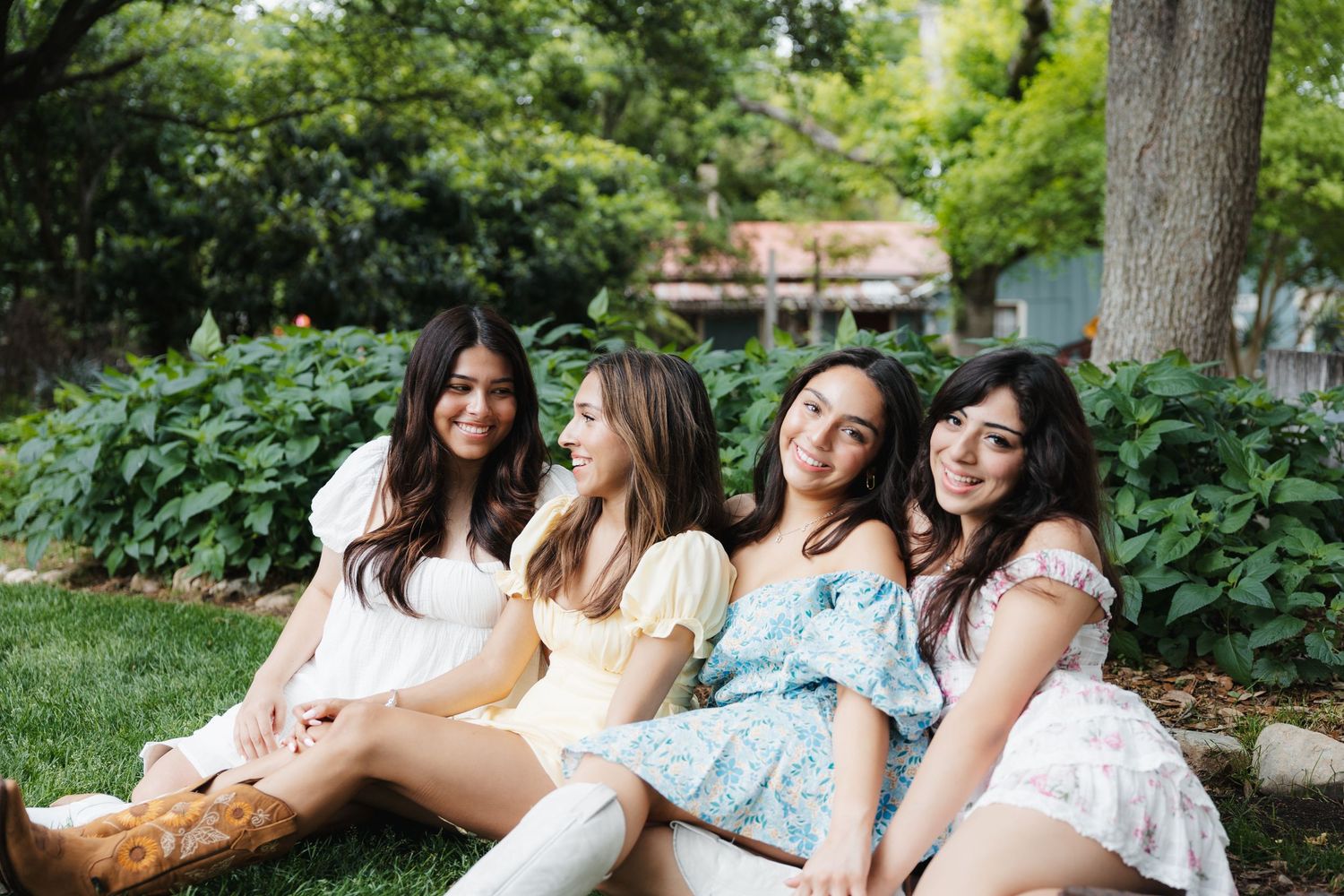 Four friends in summery dresses sit together and laugh on grass in a lush garden setting.