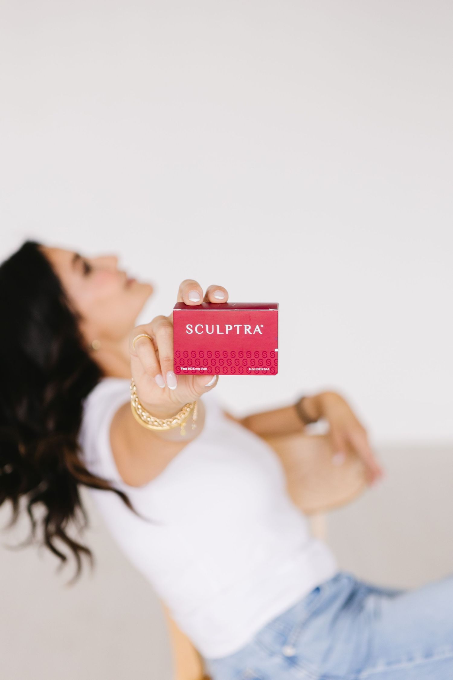 Woman in white tee holding a red Sculptra card toward camera, wearing a gold bracelet, light background.