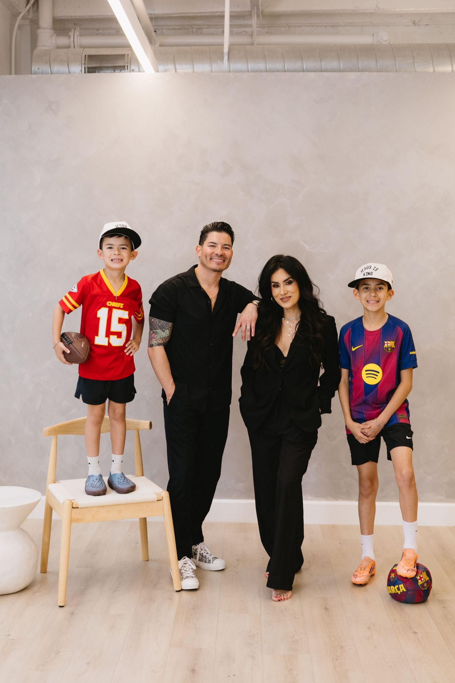Family of four posing together in a bright studio, two boys in sports jerseys holding a football and soccer ball.