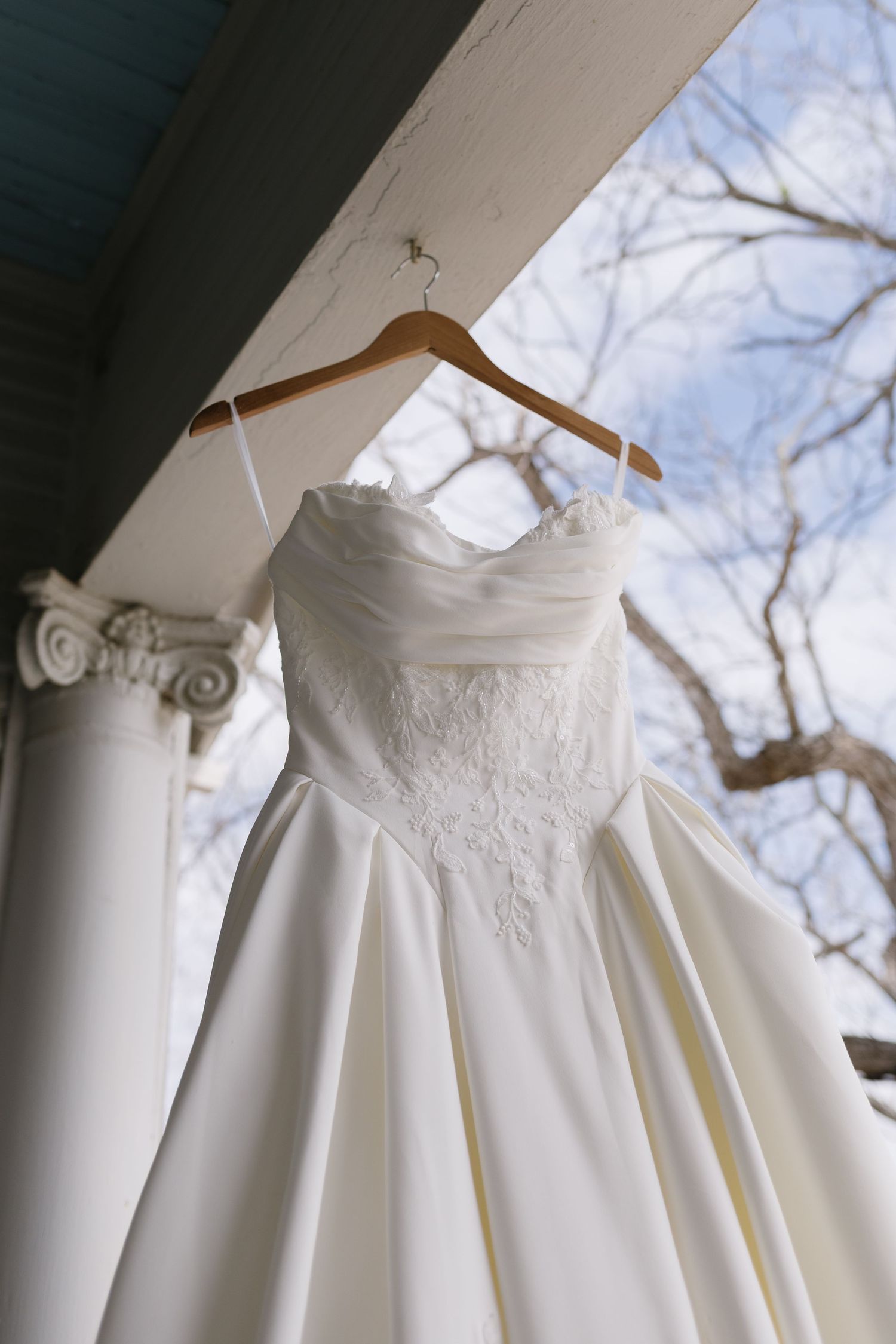 Elegant strapless white wedding gown with lace bodice hanging on wooden hanger on porch column.