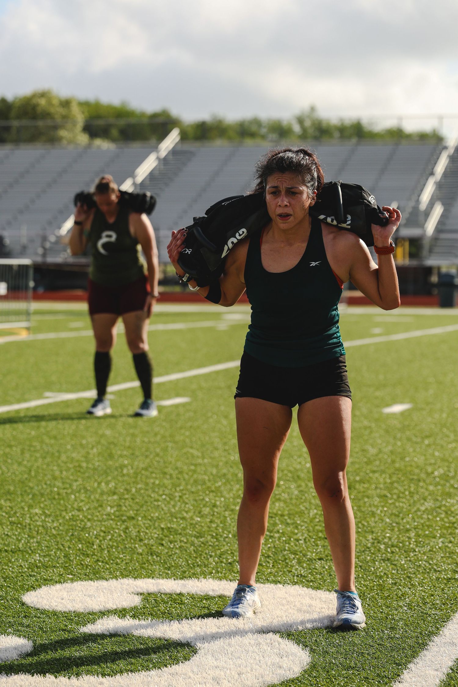 Two female athletes carry weighted sandbags on their shoulders during an outdoor training session on a football field.