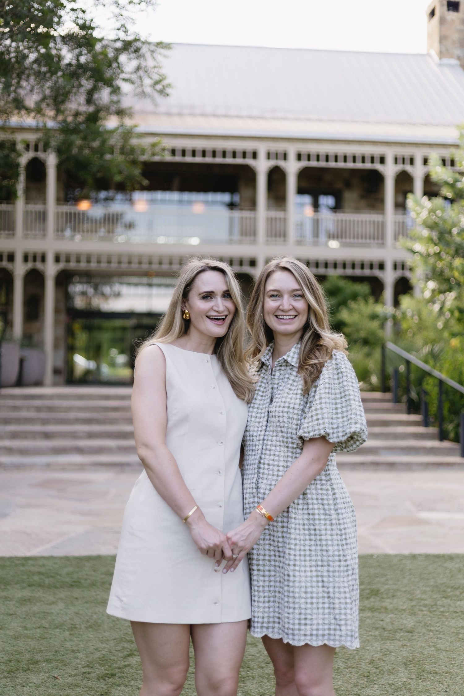 Two smiling women in summer dresses pose together outdoors near a historic building with wraparound porch.