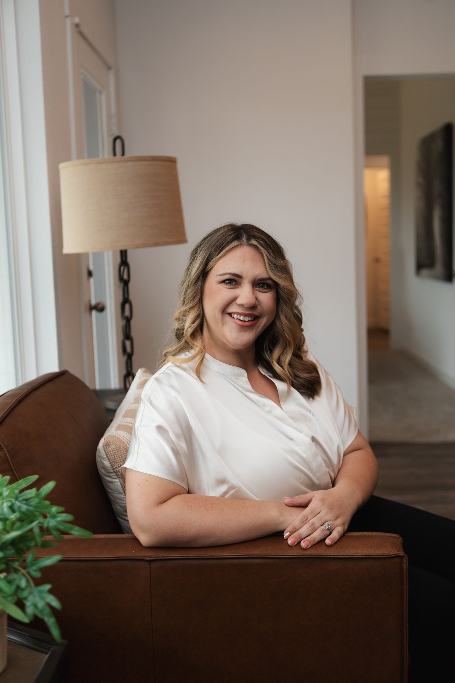 A person sits on a brown couch wearing a white blouse, with a floor lamp and indoor plant visible in the background.
