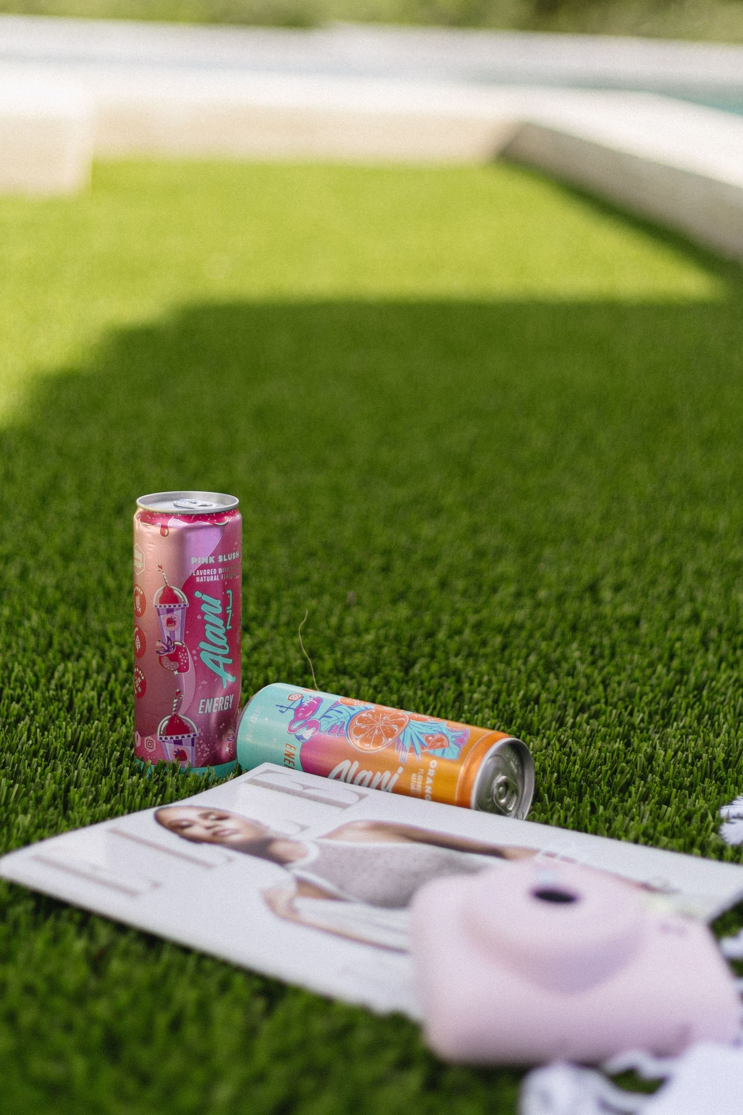 Two Alani Nu energy drink cans on a magazine and grass near a pool on a sunny day.