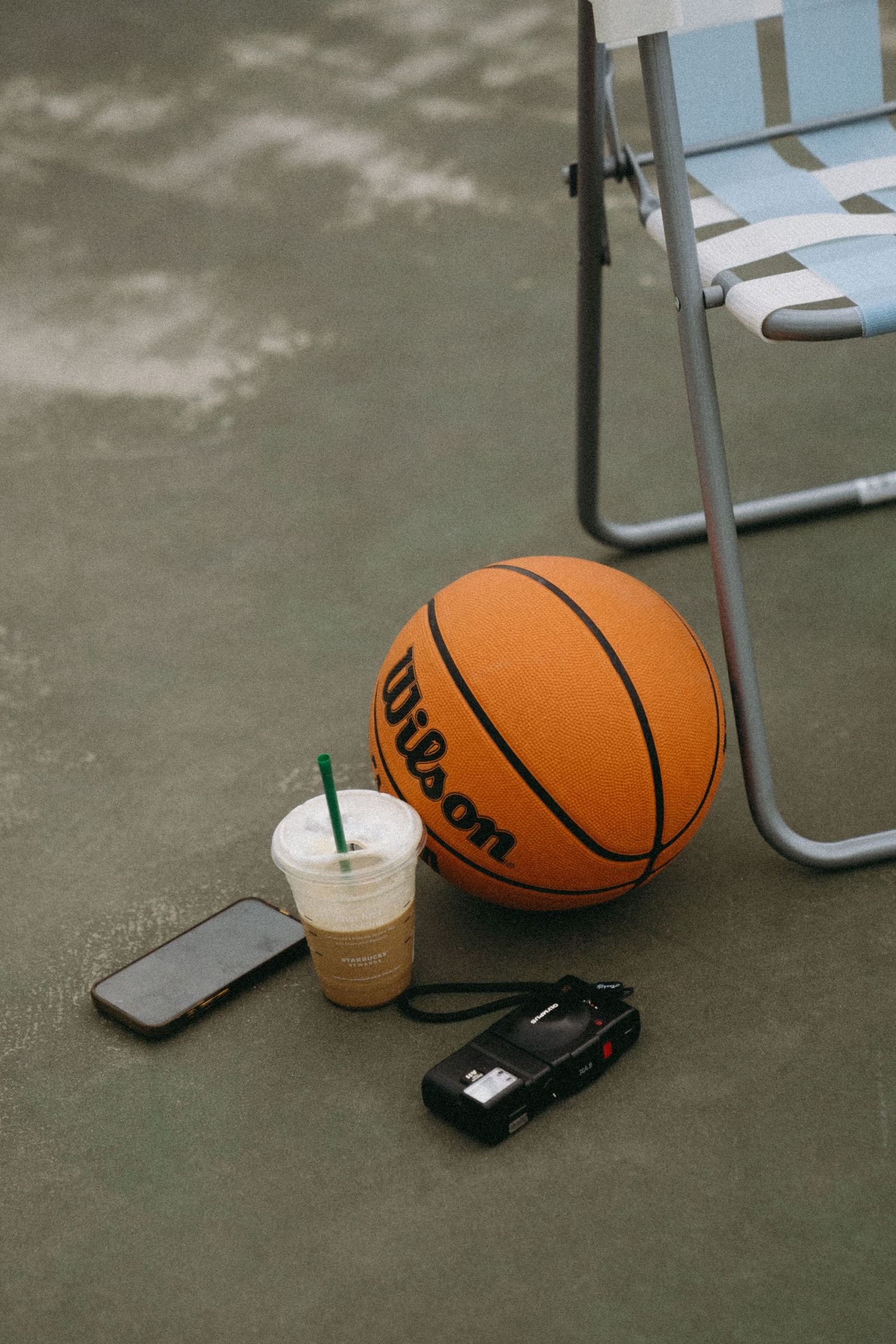 Wilson basketball beside an iced Starbucks coffee, smartphone, and camera on concrete floor near a folding chair.