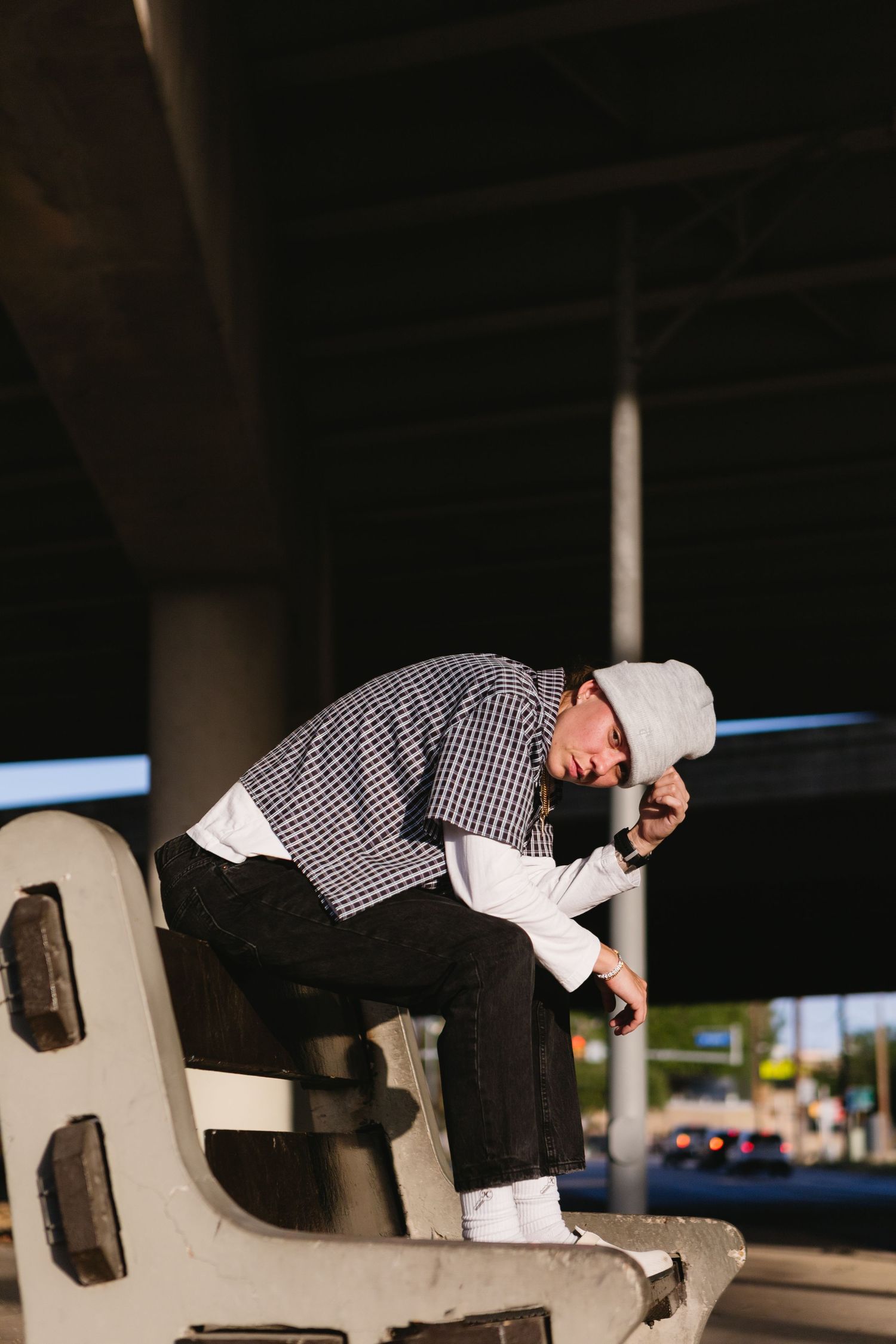 Young man in plaid shirt and white cap sits on concrete barrier under urban overpass, looking down at phone.