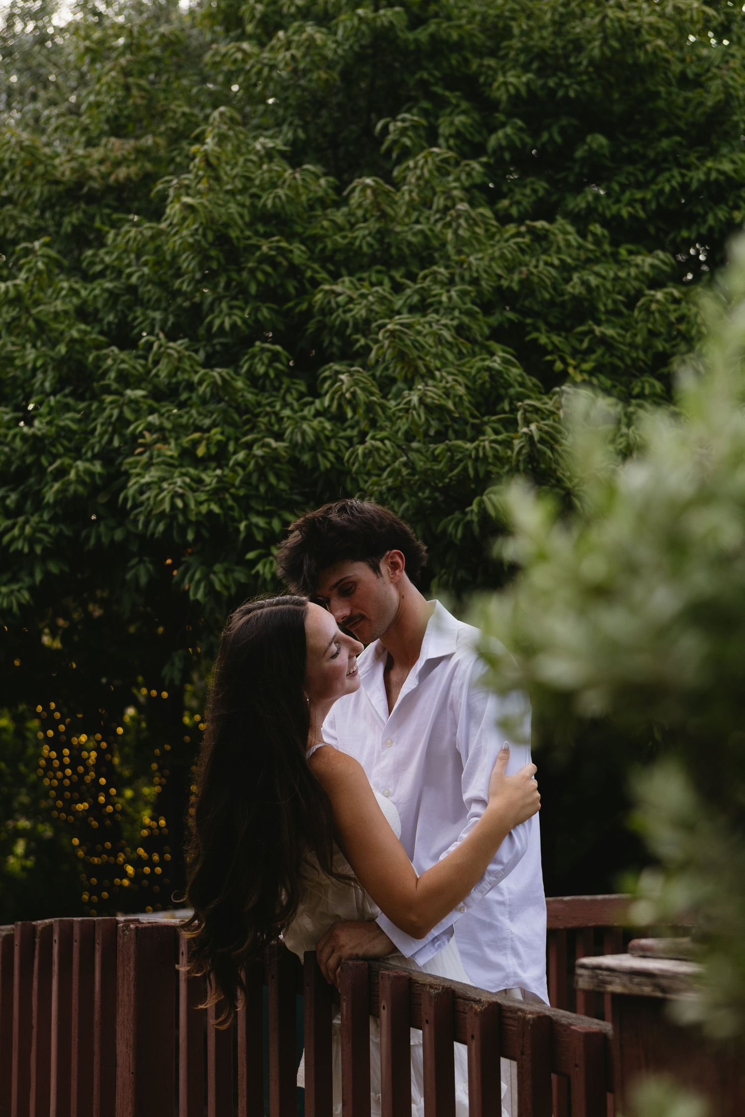 A young couple shares a romantic embrace on a wooden bridge surrounded by lush green trees.