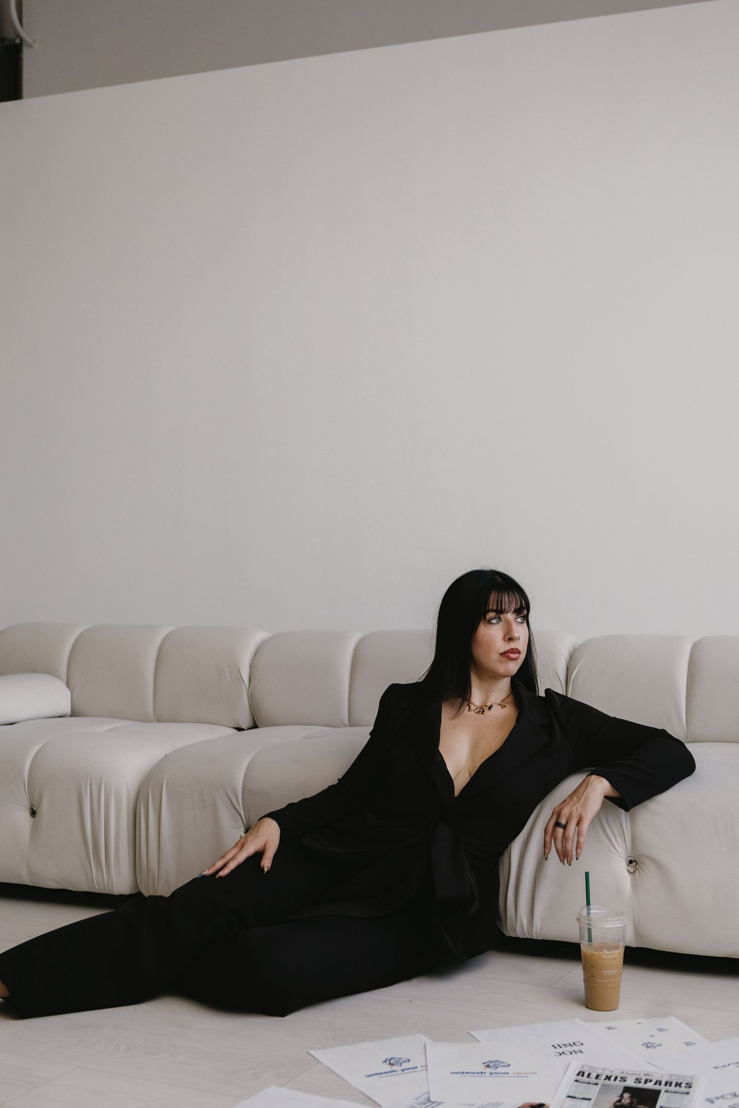 Woman in black suit lounging on white floor beside cream sofa with iced coffee nearby.