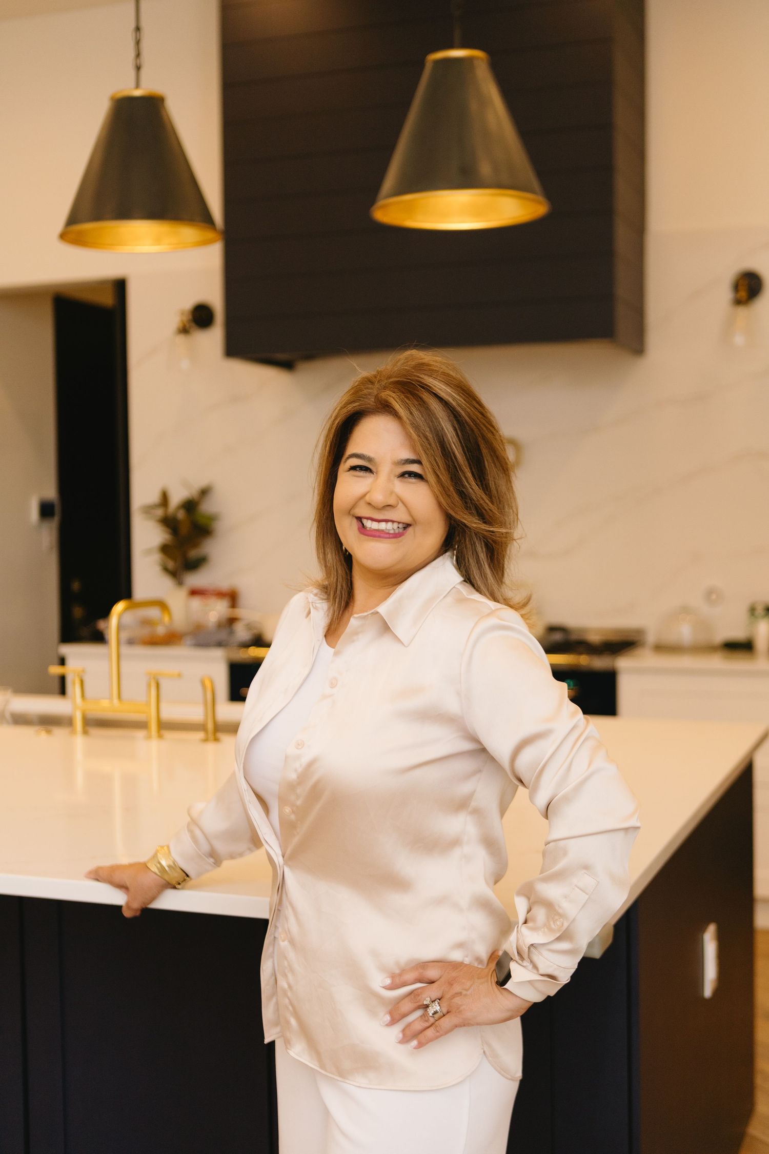 Smiling woman in satin blouse poses confidently in modern kitchen with black cabinets and gold pendant lights.