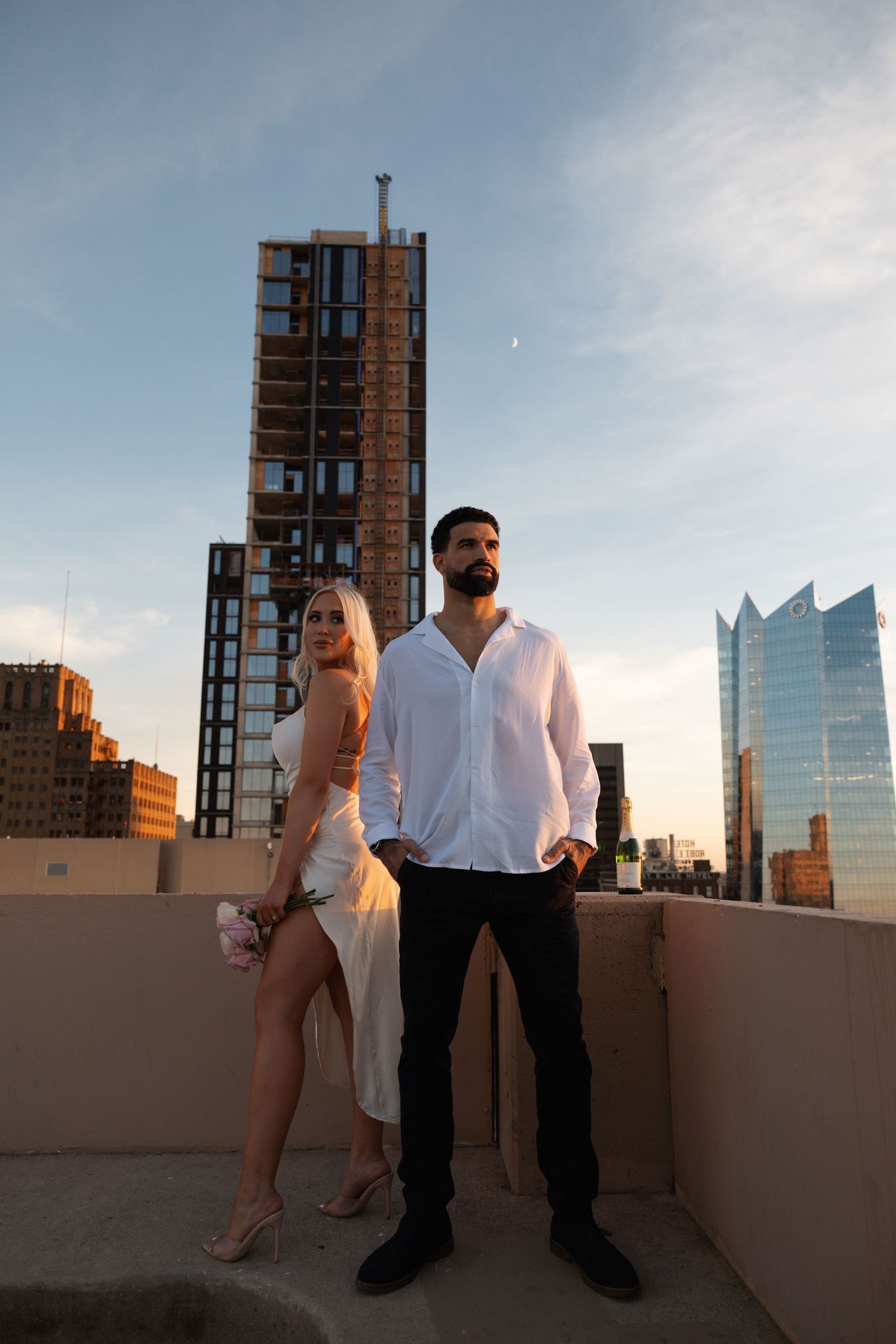 A couple poses against the skyline of a modern city at dusk.