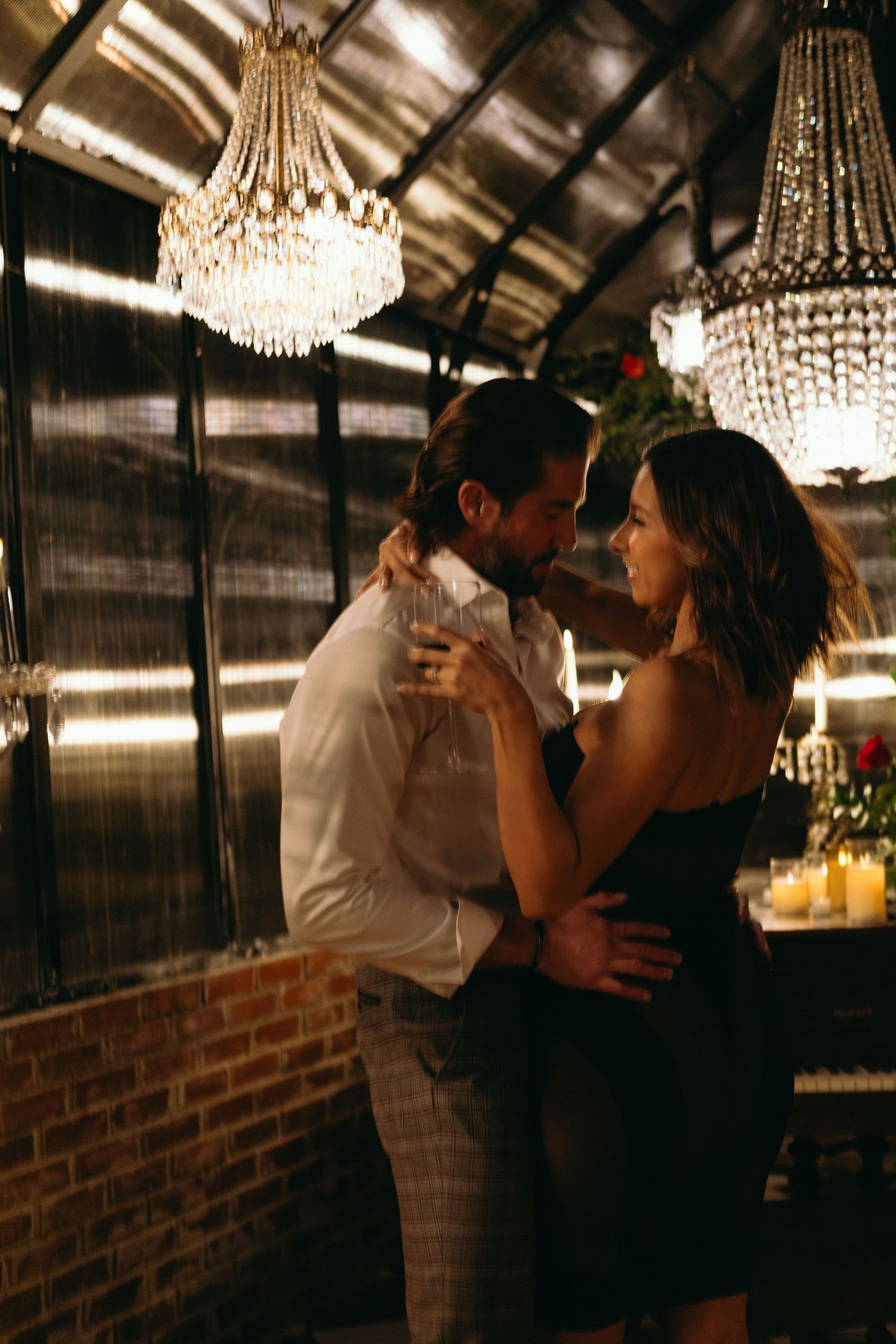 Couple dancing intimately under crystal chandeliers in a warmly lit romantic venue with brick walls.