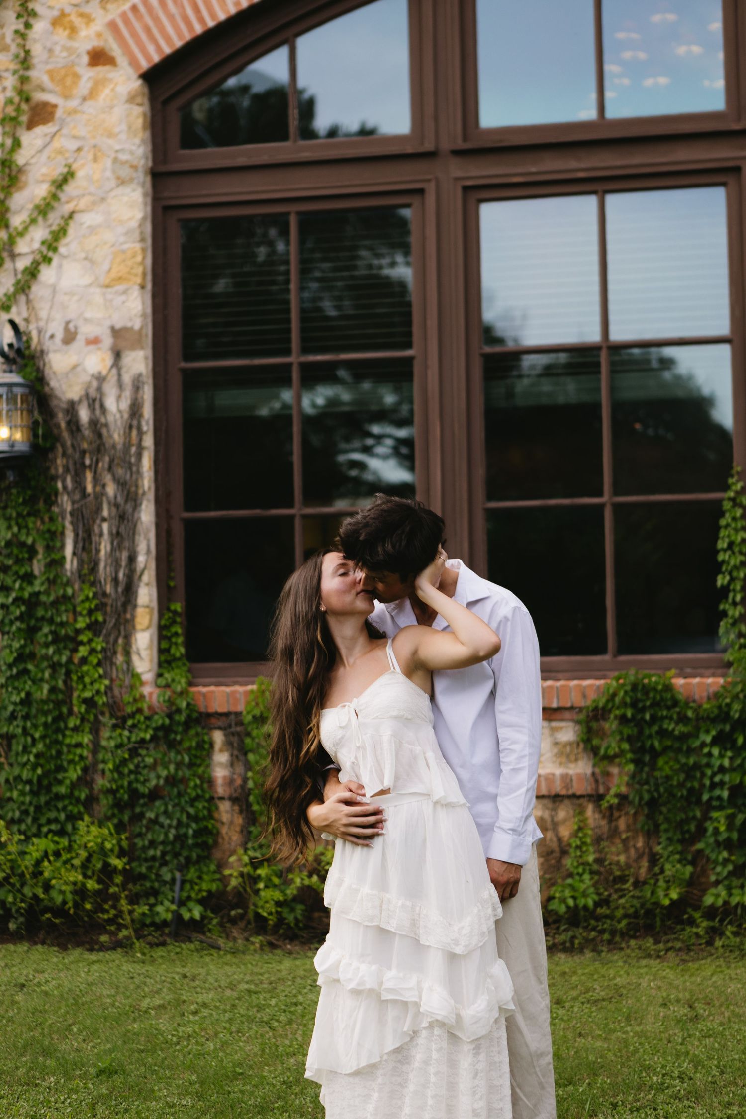 A couple shares a romantic kiss in front of a stone building with large windows and ivy, bride in white ruffled dress.