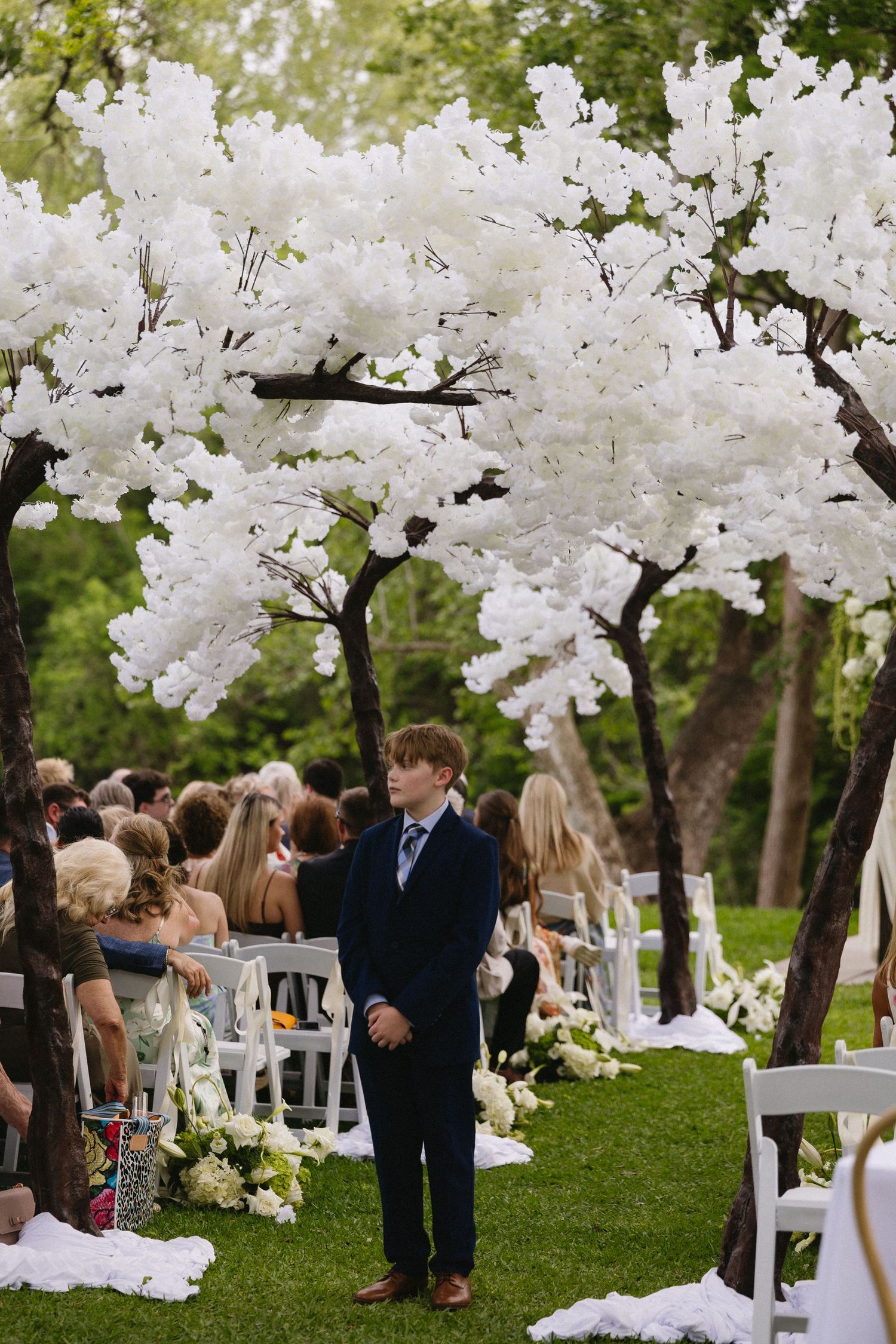 Young boy in navy suit standing at outdoor wedding ceremony aisle lined with white blossom trees and floral arrangements.