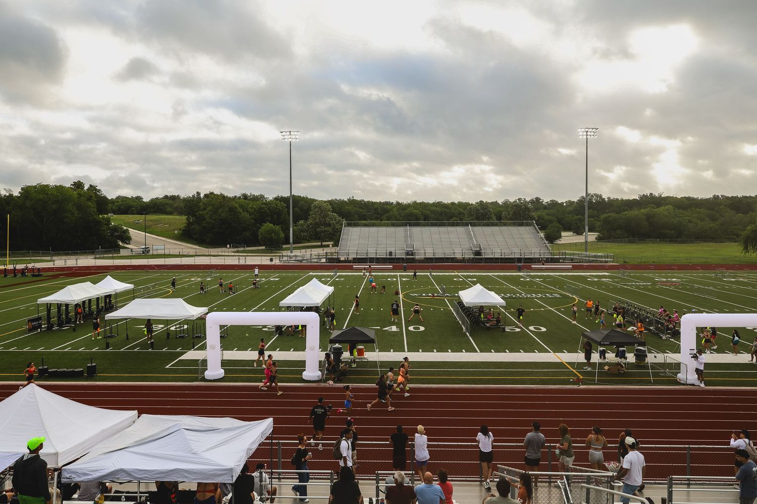 Aerial view of a football field with white inflatable arches, red track, and bleachers under a cloudy sky.