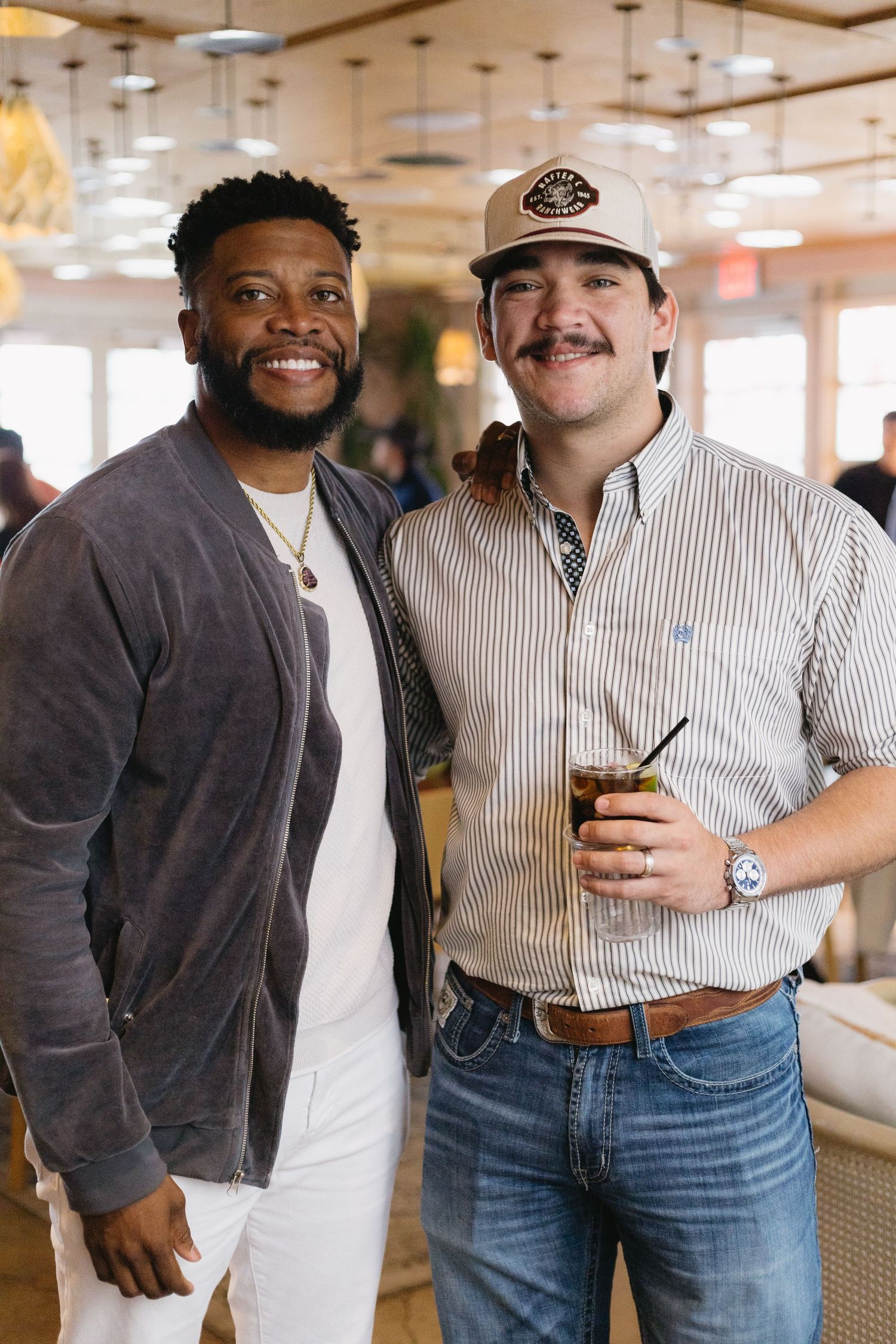 Two smiling men pose together at a restaurant, one holding a cocktail drink.