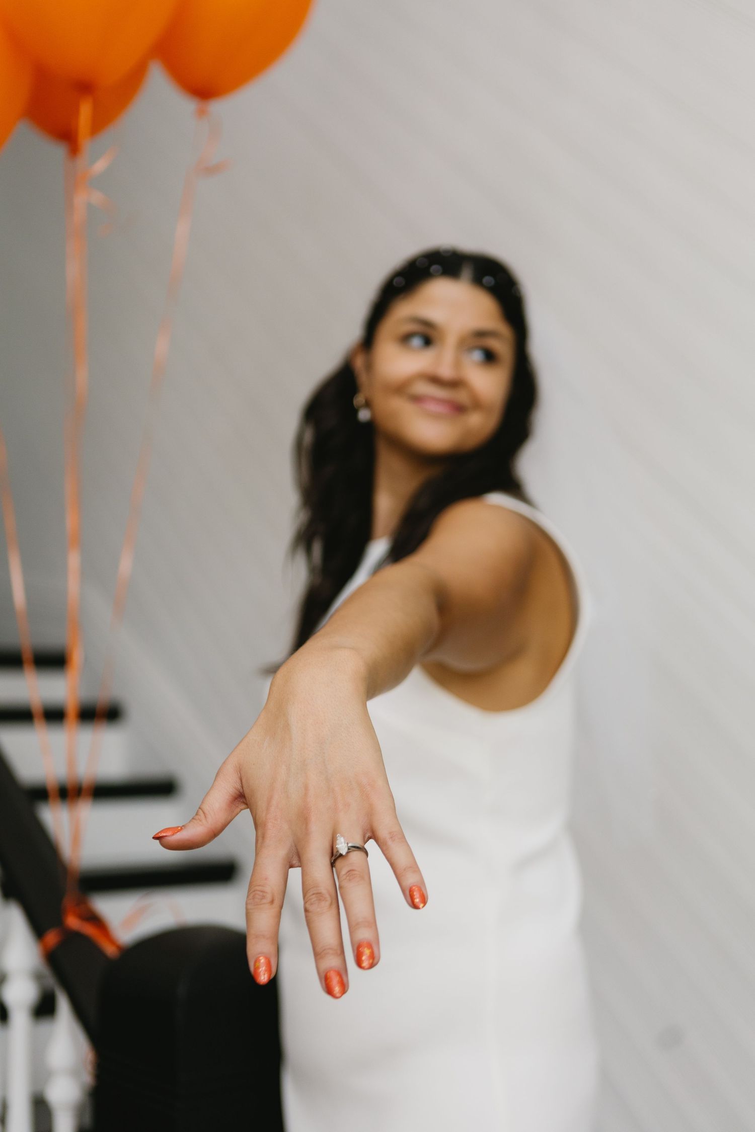 Smiling bride in white dress showing off engagement ring with orange balloons in background.