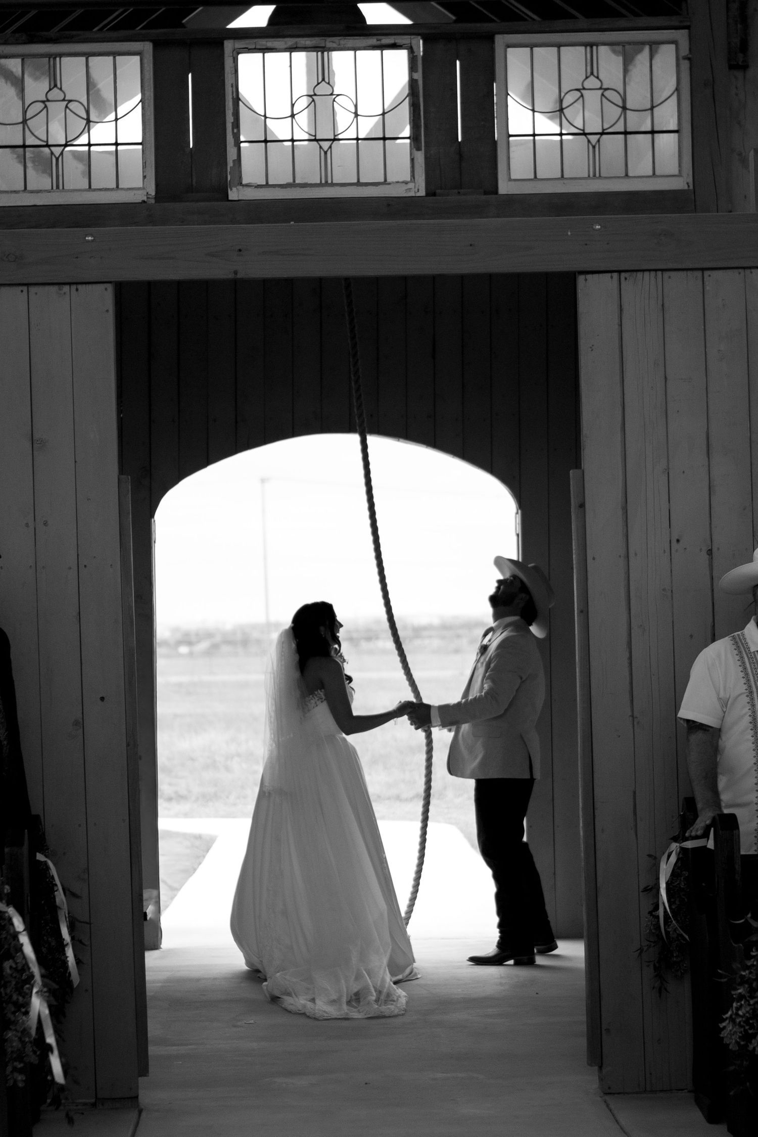 Black and white photo of bride and groom holding a rope in barn doorway at rustic country wedding.