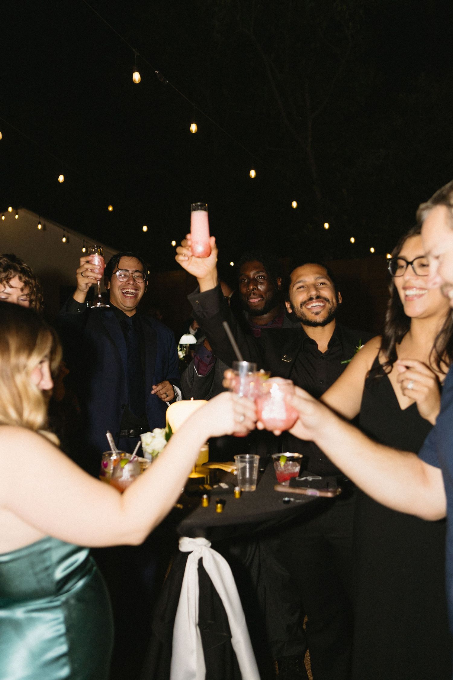 Group of friends toasting drinks at an outdoor evening party with string lights in the background.