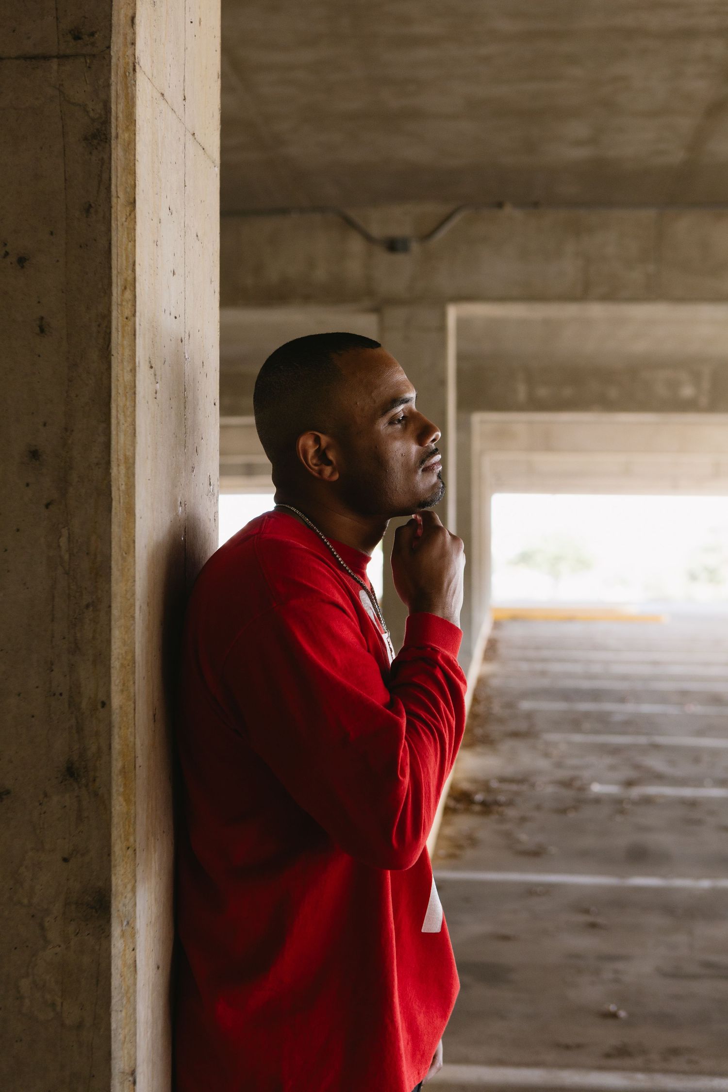 A thoughtful young Black man in a red sweatshirt leans against a concrete pillar in a parking garage, gazing upward.