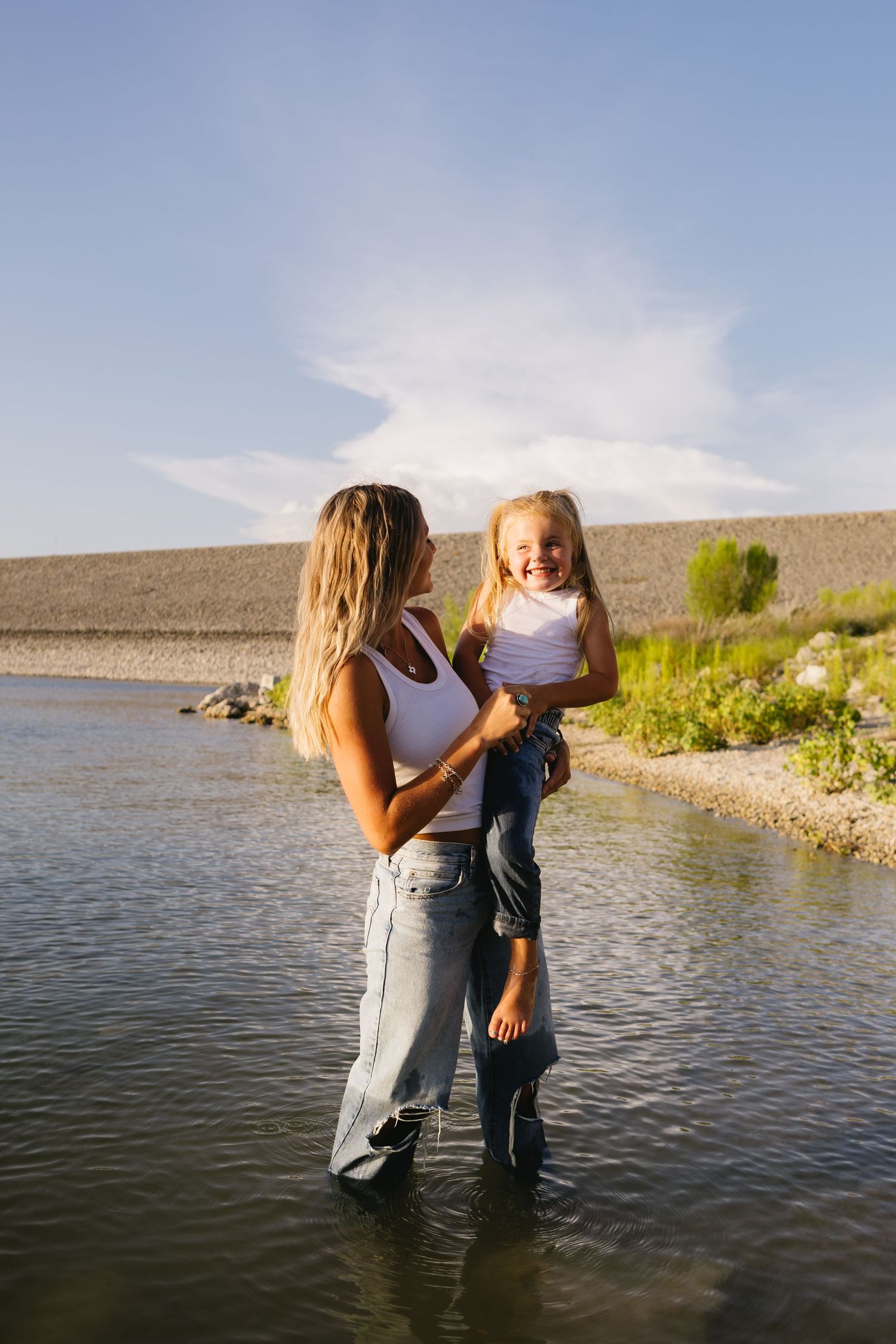 A woman stands in a shallow lake holding a laughing young girl, both wearing white tops and jeans on a sunny day.