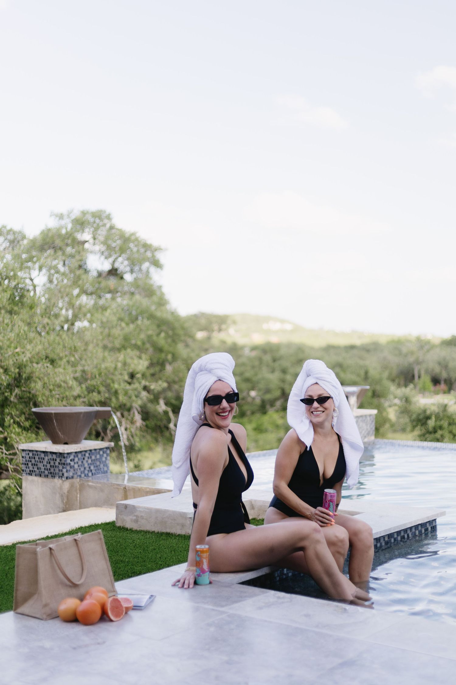 Two friends in matching black swimsuits and towel turbans lounging by luxury infinity pool with snacks.
