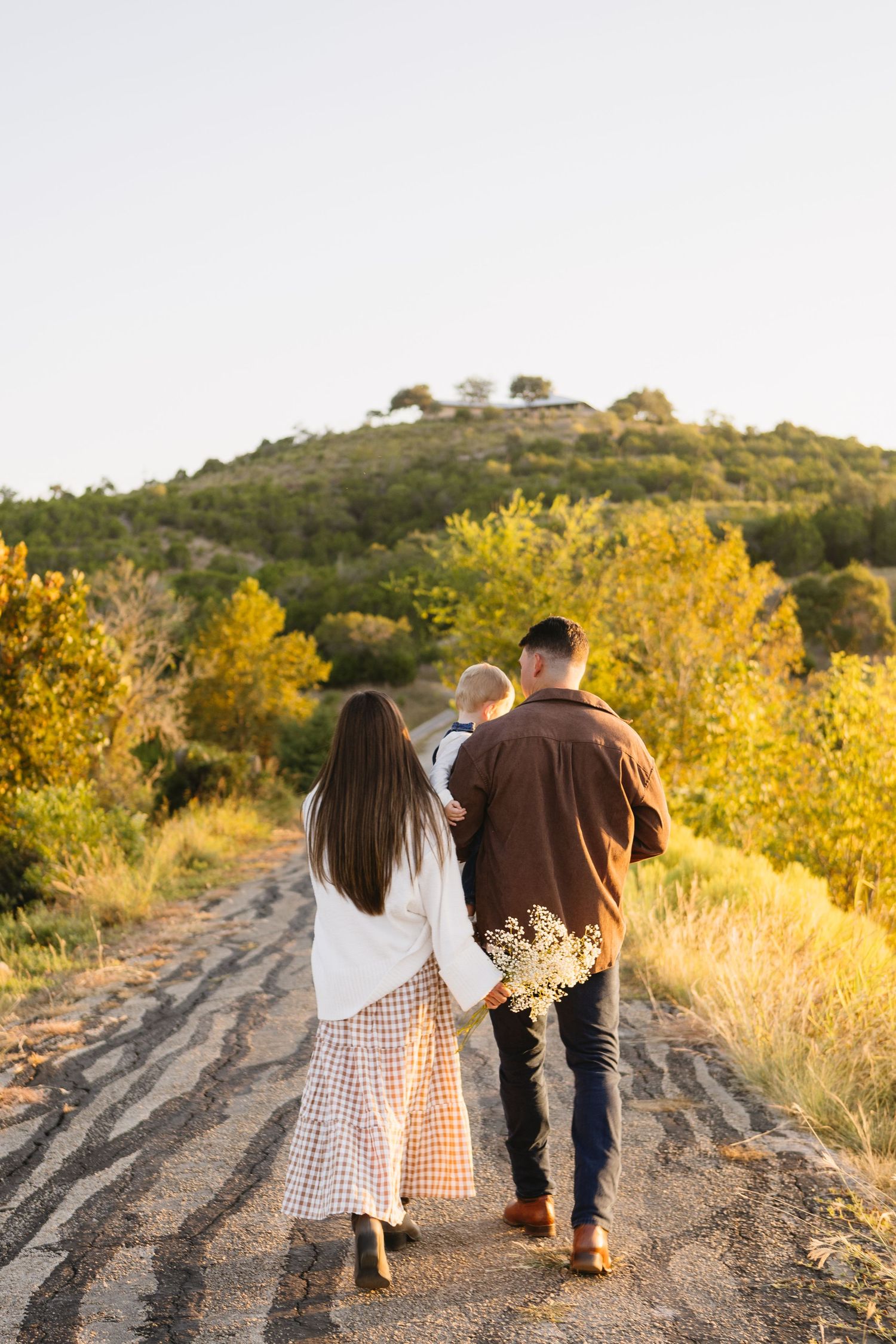 Family walking down a rural dirt road surrounded by golden autumn trees and rolling green hills at sunset.