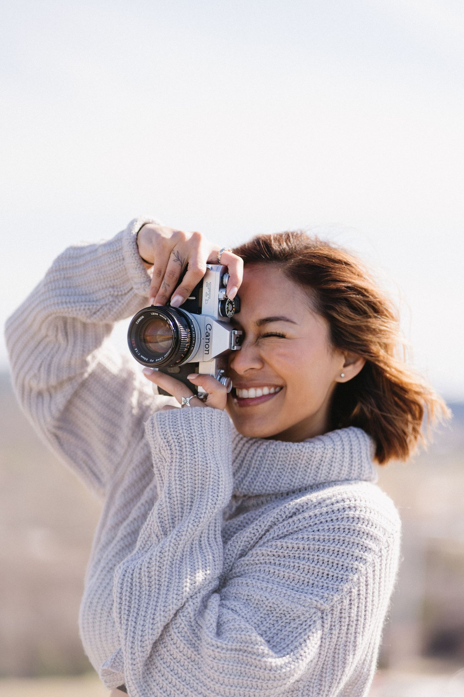Smiling woman in cozy knit sweater taking photos with a vintage film camera outdoors in natural light.