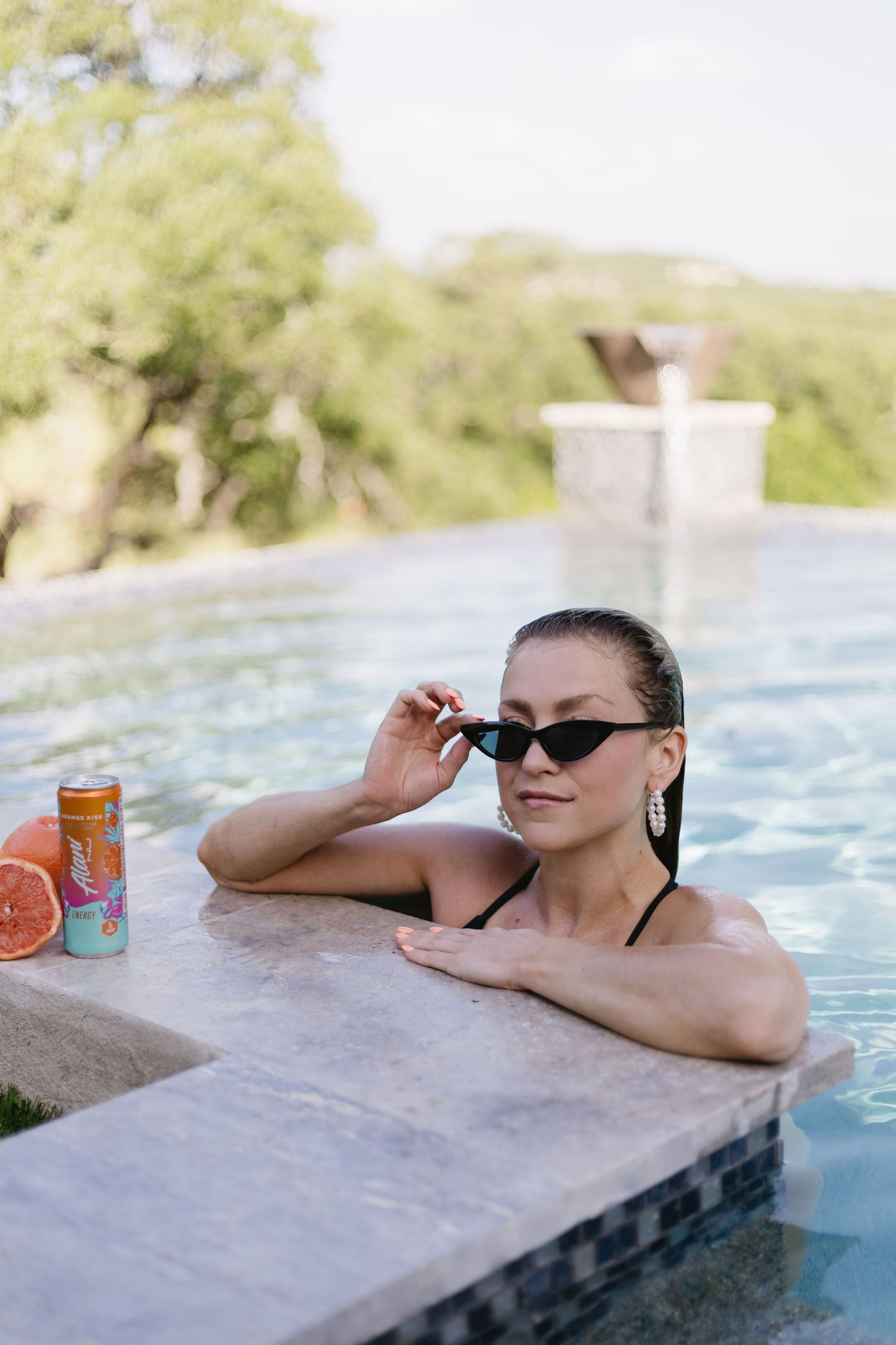 Woman in black bikini and cat-eye sunglasses relaxing in infinity pool with a canned drink on the ledge.