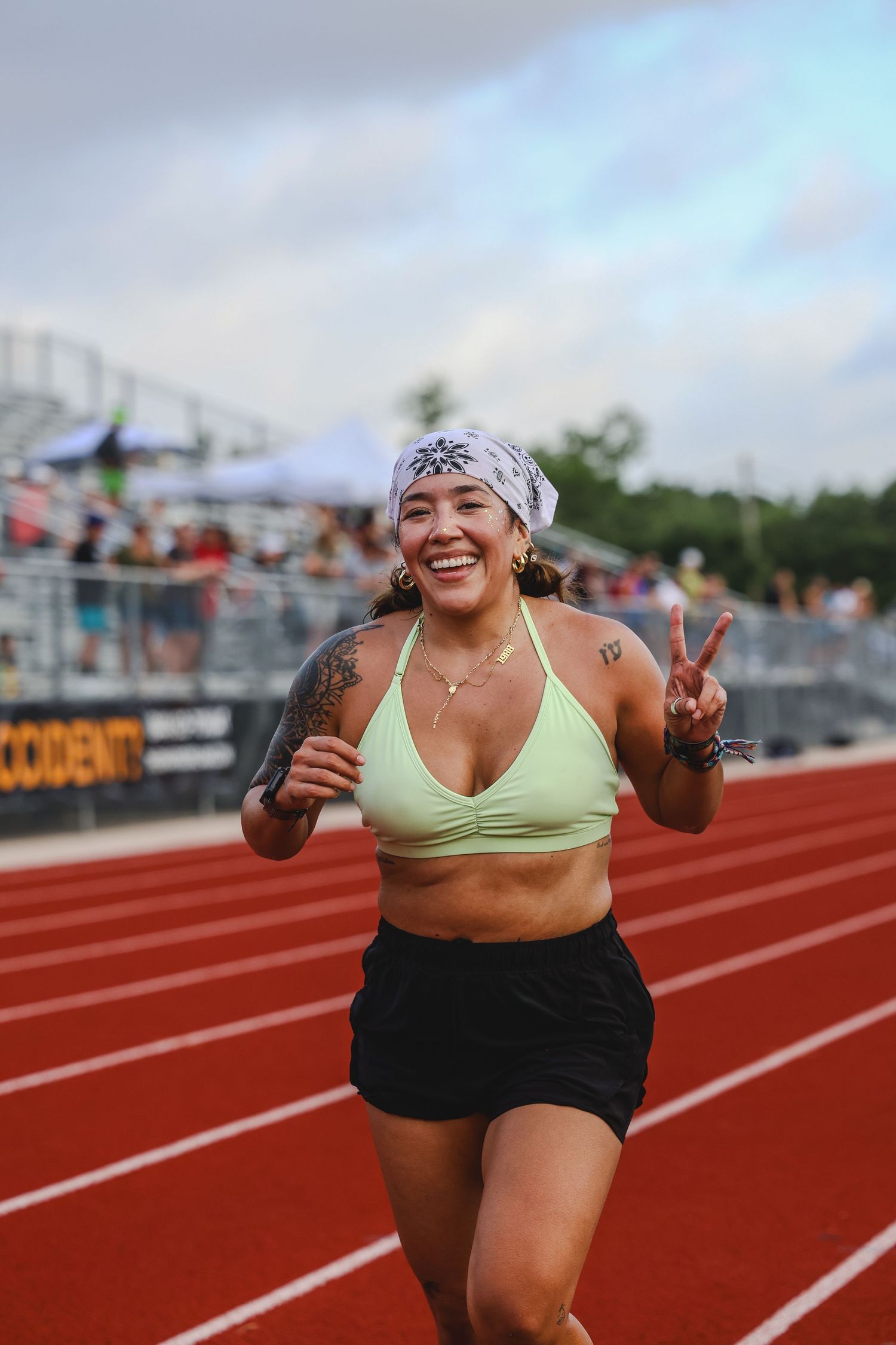 Smiling woman in green sports bra and bandana flashing peace sign while running on red track.