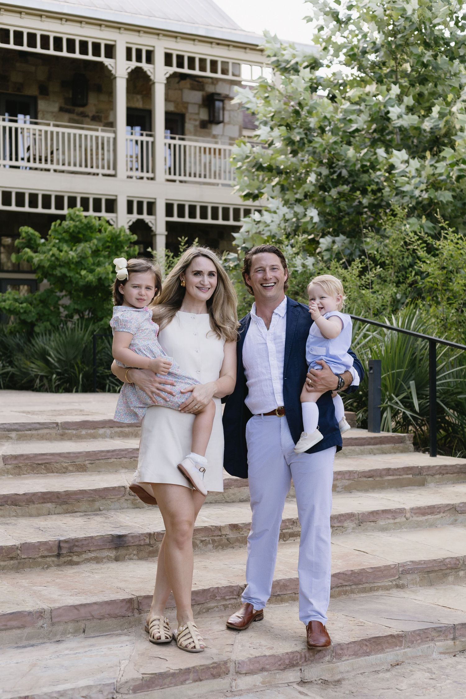 A smiling family of four poses on stone steps, parents holding two young children, with a historic building behind them.