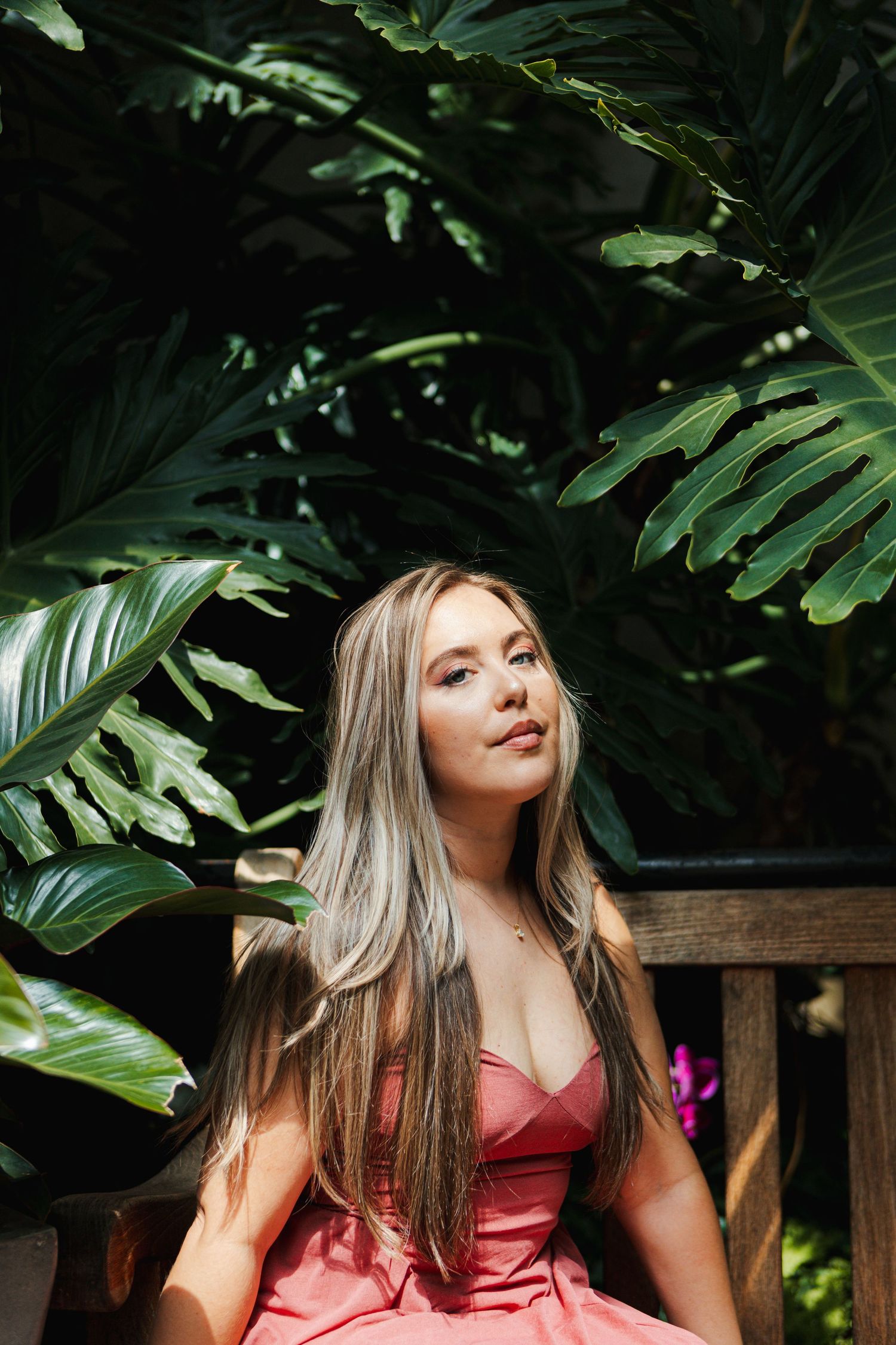 Portrait of someone with blonde hair wearing a red dress posing outdoors among tropical foliage and garden bench.