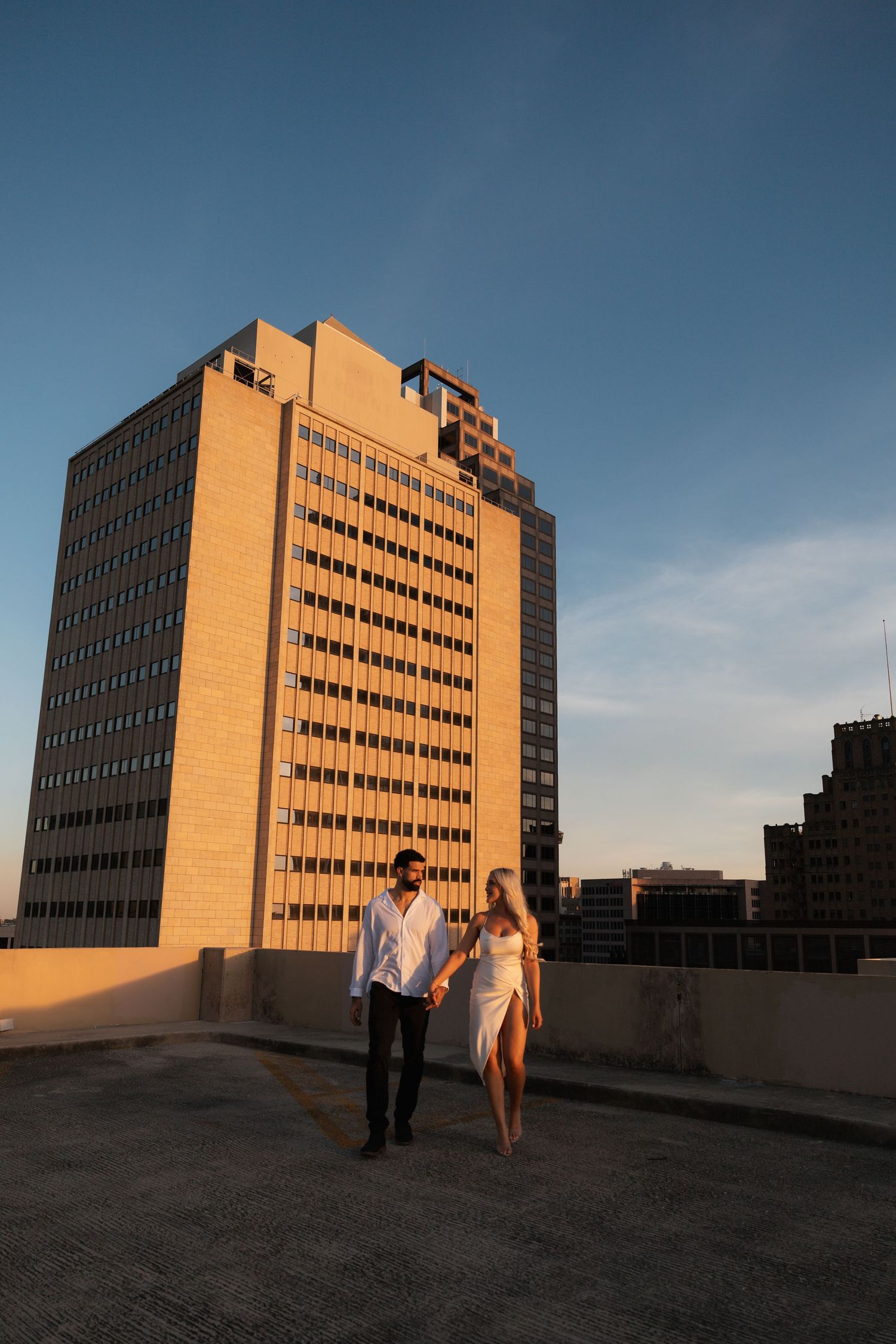 A couple walks along a rooftop with golden sunlight illuminating tall buildings.