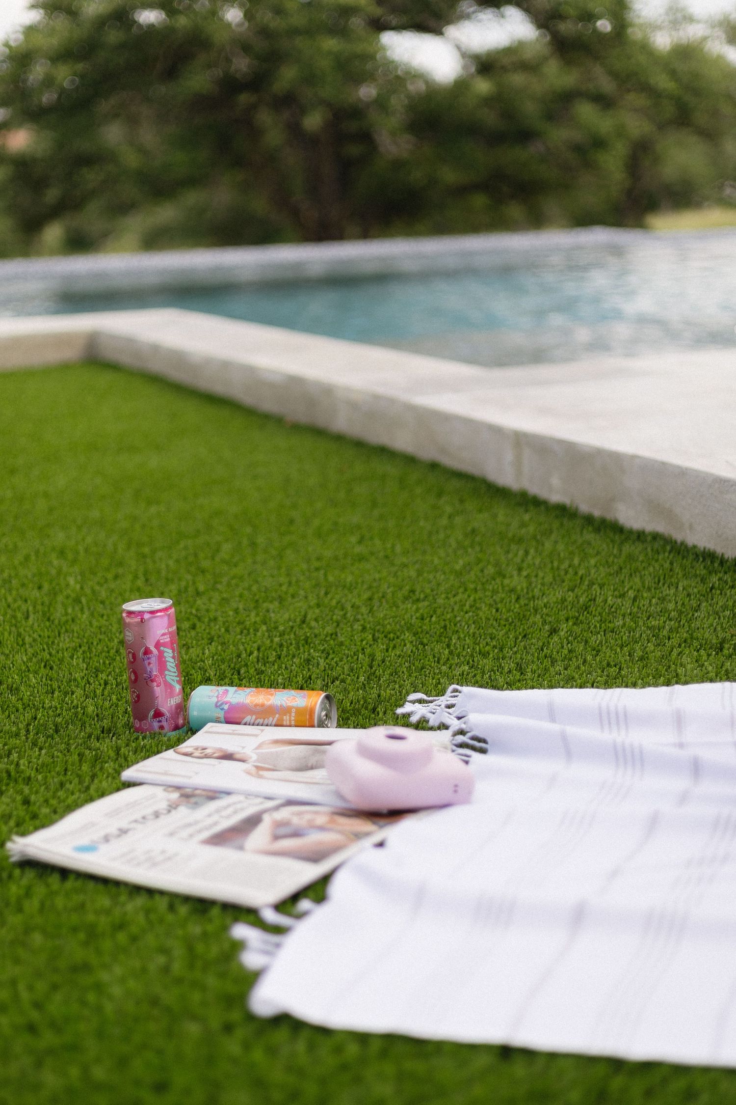 White towels, magazines and drinks laid out on artificial grass next to a modern swimming pool.