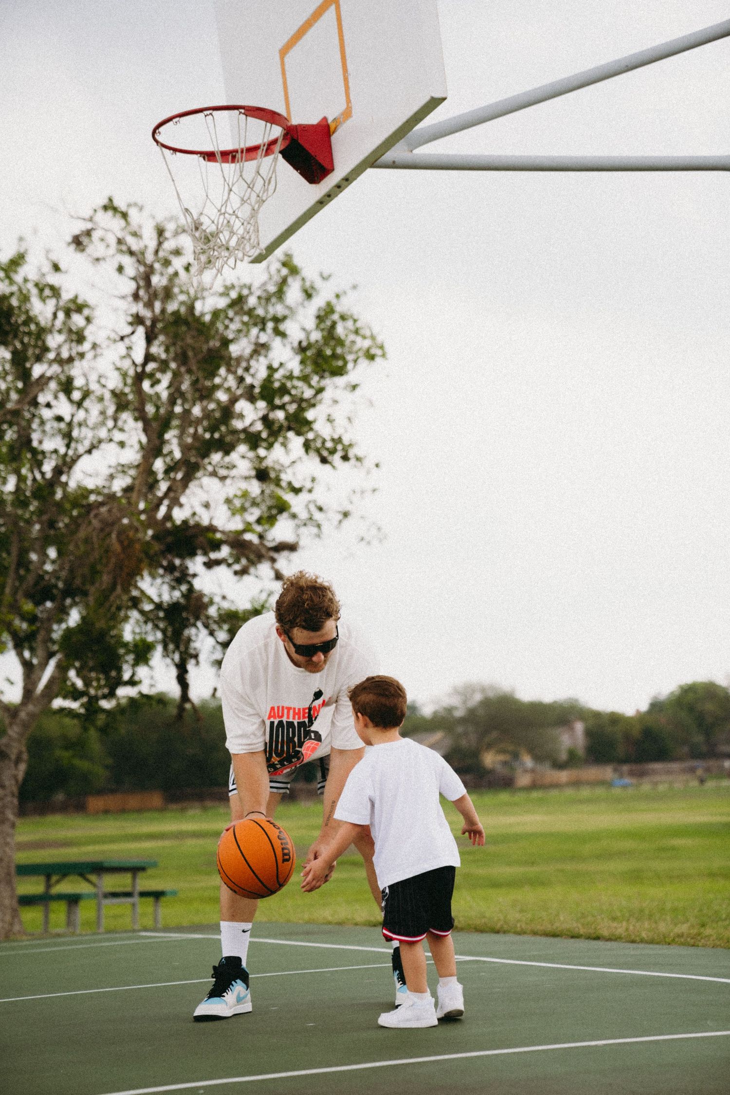 Man in Jordan shirt teaching young boy basketball on outdoor court.