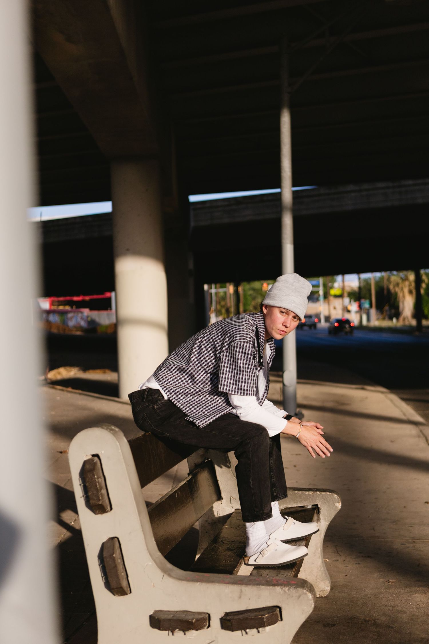 Young man in plaid shirt and gray beanie sits on concrete bench under urban overpass.
