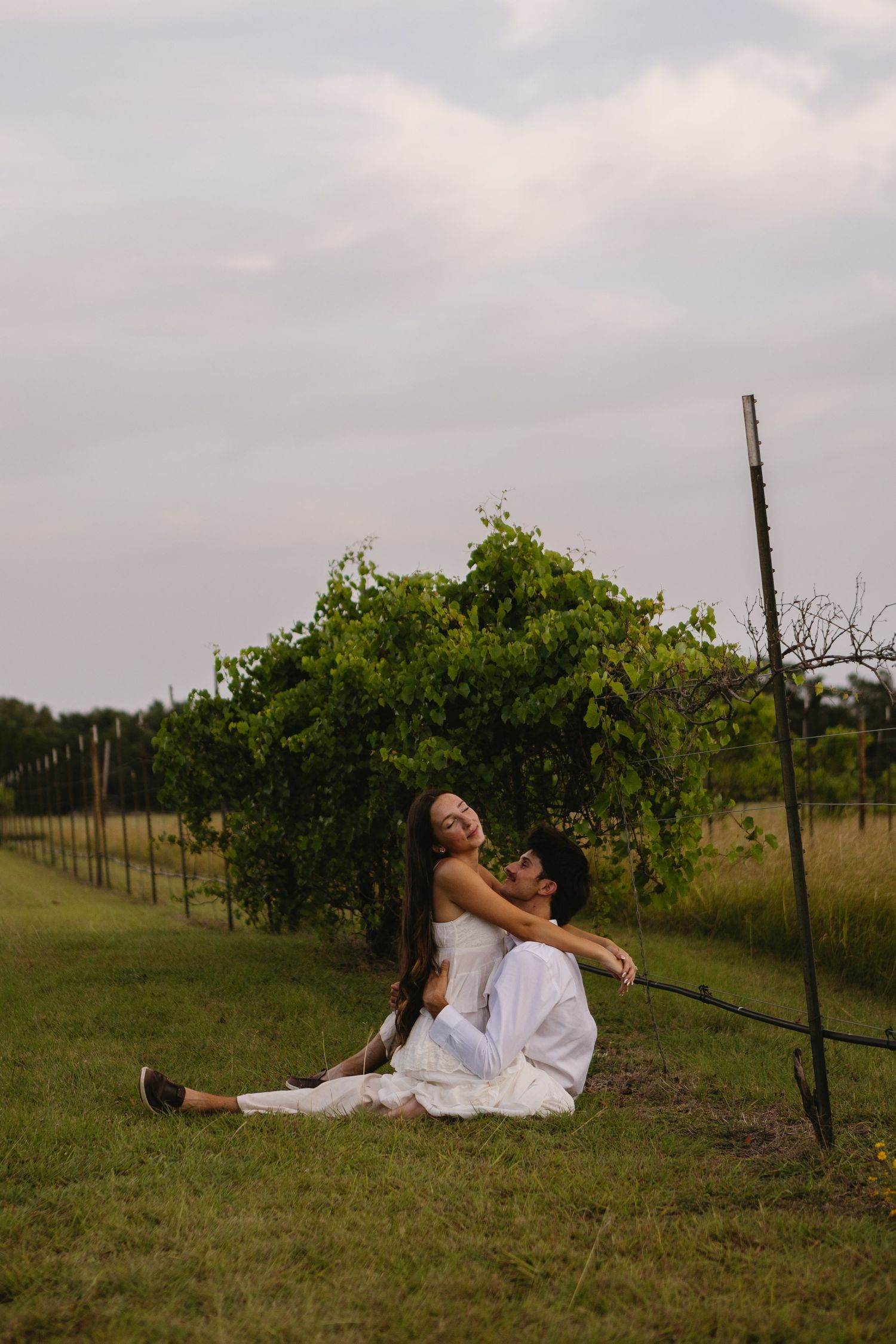 Couple in white sitting on grass at a vineyard, woman laughing joyfully as man holds her in romantic outdoor setting.