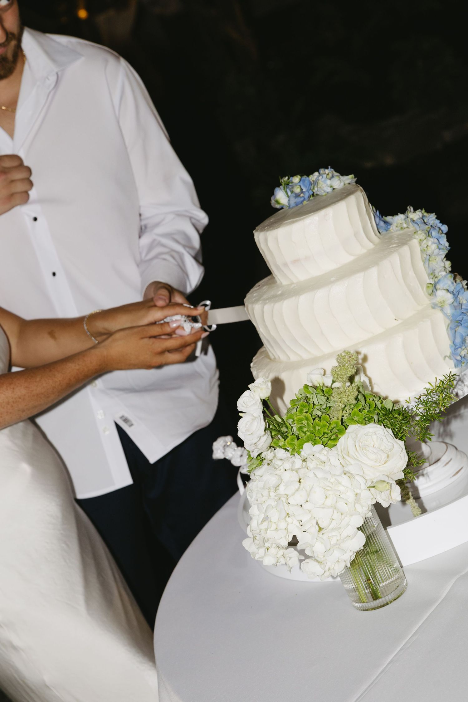 Couple cutting a three-tier white wedding cake adorned with blue hydrangeas and white roses at evening reception.