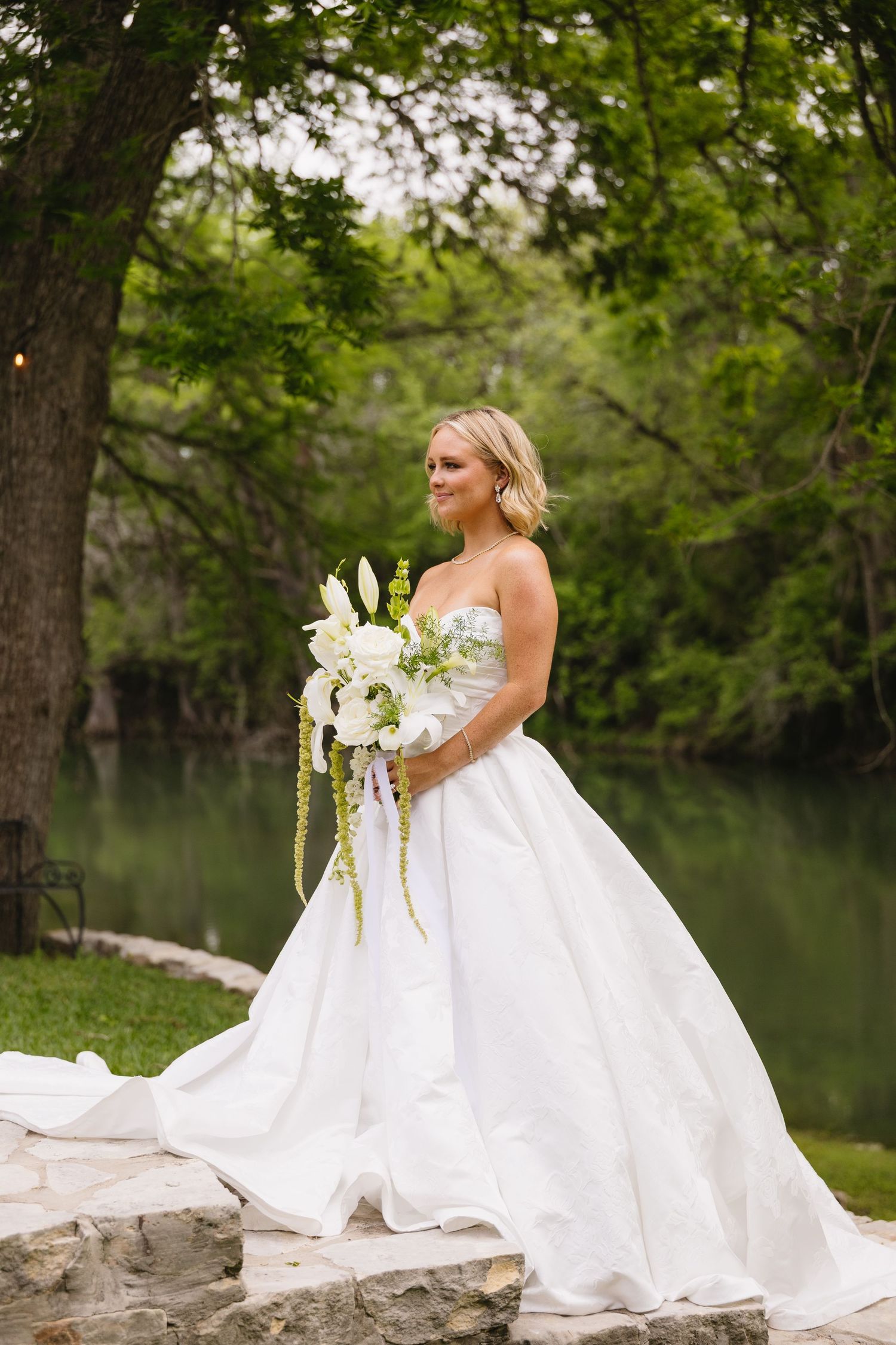 Bride in strapless white ball gown holding cascading white floral bouquet standing on stone steps by a serene river.