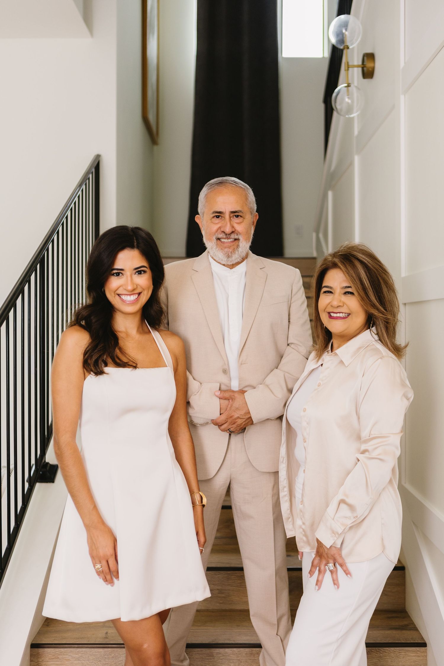 Three smiling people in white and beige outfits pose together on a staircase in a modern home.